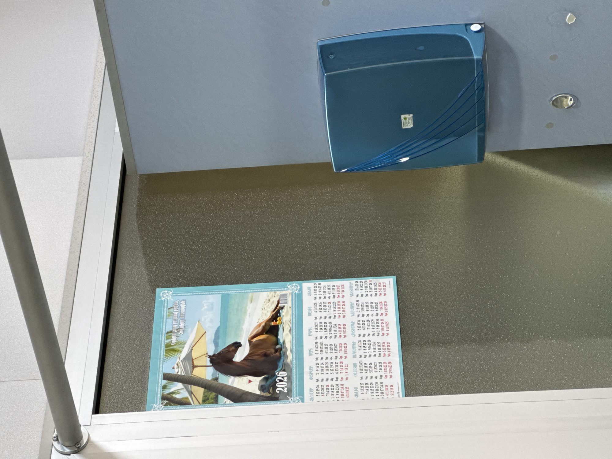 A straight-on shot presents a calendar displaying a horse illustration and a blue rectangular container mounted on a speckled wall. The calendar, positioned below the container on a surface (possibly a shelf or step), features the year "2026" beneath the horse picture and includes Armenian text above. The wall itself has a rough texture. Above the wall is a flat, bluish-gray ceiling with mounted hardware. A gray bar descends vertically on the left side of the frame, suggesting a structural element. The container appears to be made of shiny blue plastic. The location is unknown, Yerevan is not discernible from the image.