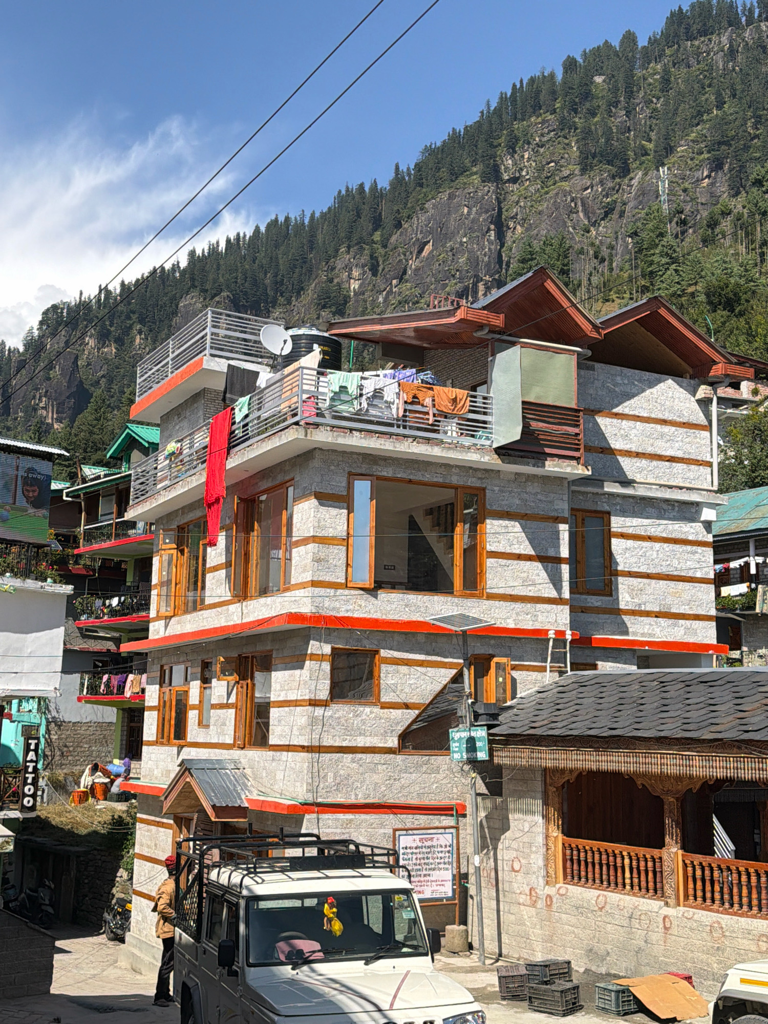 The image depicts a street scene in Vashisht, India, with a multi-story building dominating the foreground. The building features a stone facade with wooden accents and red trim. Laundry is visible drying on balconies, and a satellite dish is mounted on the roof. A white SUV is parked on the street, with a person in a brown jacket standing next to it. In the background, a steep, tree-covered mountainside rises under a blue sky with scattered clouds. Signs are visible on some of the buildings, including one that says "TATTOO" and another with Hindi text and "NO SMOKING." The overall environment suggests a residential or commercial area nestled in a mountainous region.