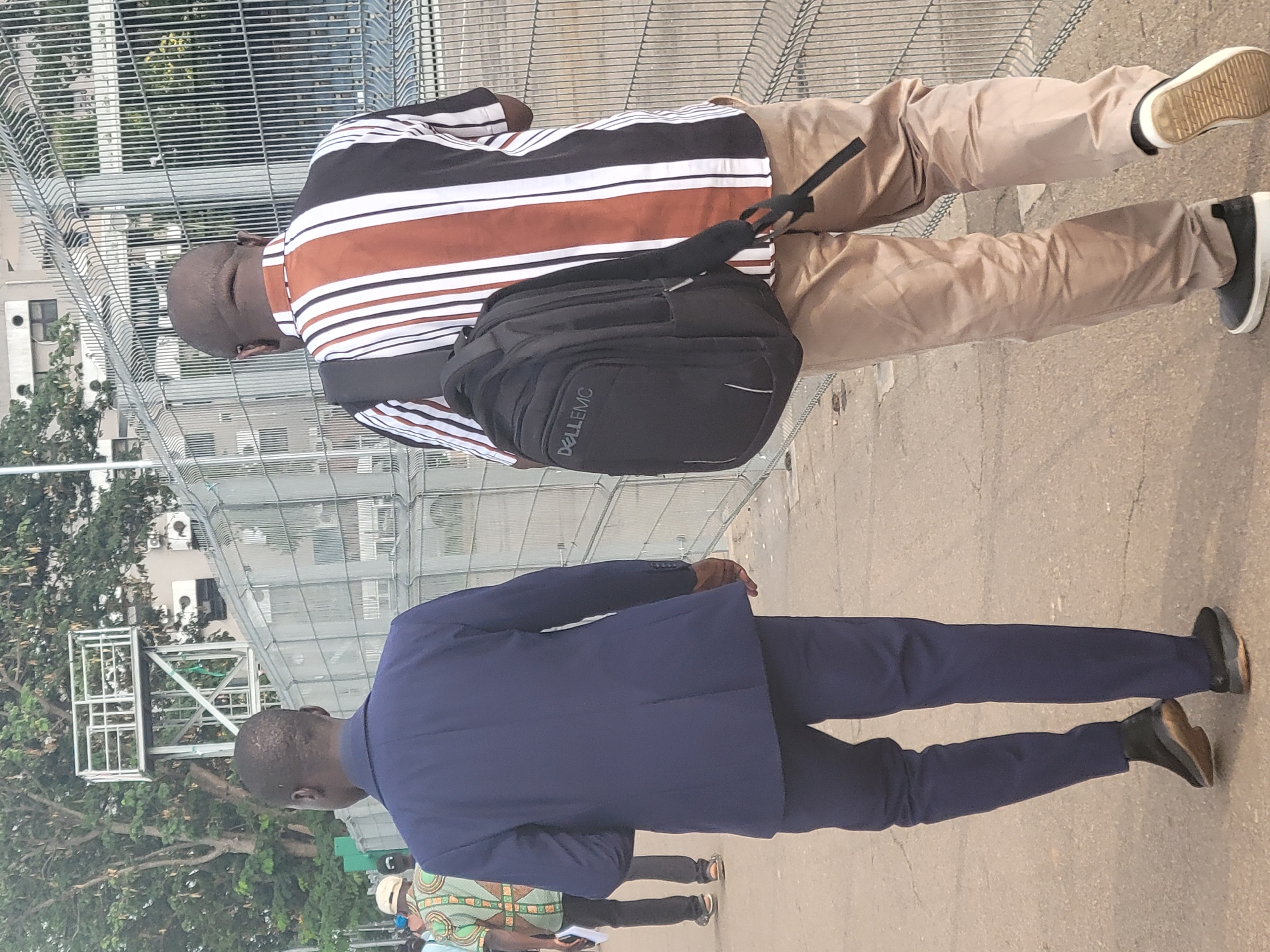 An outdoor image captures three individuals, primarily focusing on two men from a behind/side perspective, set against a backdrop of metal fencing, trees, and buildings in Abuja, Nigeria.
In the foreground, a man is seen from the back, wearing a dark blue suit jacket and matching trousers, paired with dark dress shoes. He has short, dark hair.
Slightly further back and to the right is another man, also viewed from behind. He is dressed in a striped polo shirt featuring horizontal bands of brown, white, and dark blue or black. He wears light brown or khaki trousers and dark casual shoes with visible white soles. A black backpack with the text "DELLEMC" clearly visible on its front pocket is worn over his right shoulder.
A third person is partially visible in the distance between the two main figures, wearing a patterned green and orange shirt.
The scene appears to be a paved area, likely a walkway or street, enclosed by prominent metal mesh fencing on either side, suggesting proximity to a secured facility or construction site. Beyond the fencing, lush green trees and modern multi-story buildings are visible under clear daylight conditions. The individuals appear to be walking or standing casually.