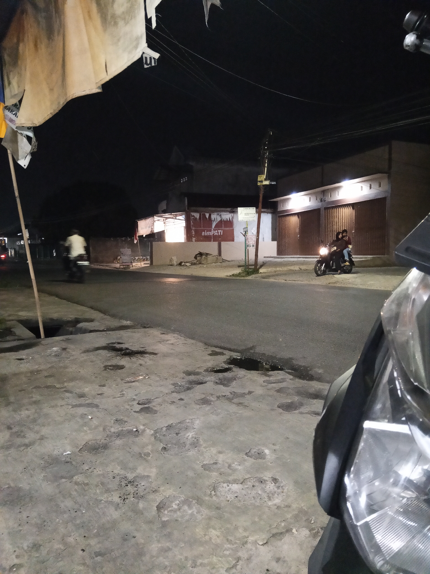 The image depicts a nighttime street scene in Lubuklinggau, Indonesia. In the foreground, the front of a dark-colored motorcycle, including its headlight, is visible. The street is paved and shows signs of wear, with oil stains and patches of darker asphalt. To the left of the street, a concrete drainage ditch and a sidewalk are visible, with a wooden pole planted in the ground.
Further down the street, a person on a motorcycle is seen in motion, blurred due to the long exposure. On the right side of the street, a building with a light-colored facade and brown roll-up doors is illuminated by overhead lights. In front of this building, another motorcycle with two occupants is parked. The passengers appear to be a woman in the front and a person in the back wearing a red shirt.
A banner with red and white imagery is visible on the building facade, and a sign with the word "SIMPATI" is also present. Overhead, power lines crisscross the dark sky. The overall atmosphere is that of a quiet, dimly lit street at night.