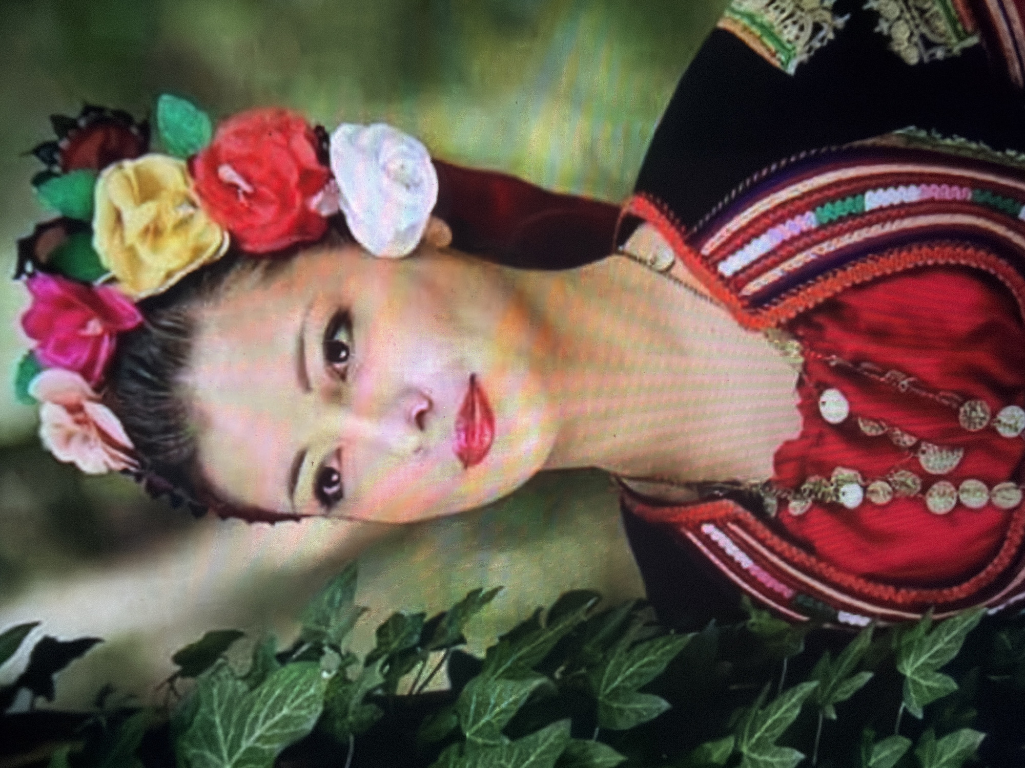A young woman is depicted in a three-quarter portrait, facing slightly to the right. She has dark hair pulled back and wears a floral headpiece composed of artificial flowers in pink, magenta, yellow, red, and white, with green foliage accents. Her face features dark eyes, defined eyebrows, and red lipstick. She is attired in a traditional Bulgarian folk costume, including a red garment underneath and a dark outer garment with a detailed, multicolored embroidered trim on the neckline and shoulders, featuring vertical stripes in various hues including white, pink, purple, and green. The visible sleeve on her right arm exhibits intricate white and gold embroidery. Multiple necklaces with round, metallic, coin-like pendants are visible around her neck. To the left of the subject, dark green ivy foliage is present. The background is a blurred green and brown, suggesting an outdoor or natural setting. The scene is located in Shumen, Bulgaria.