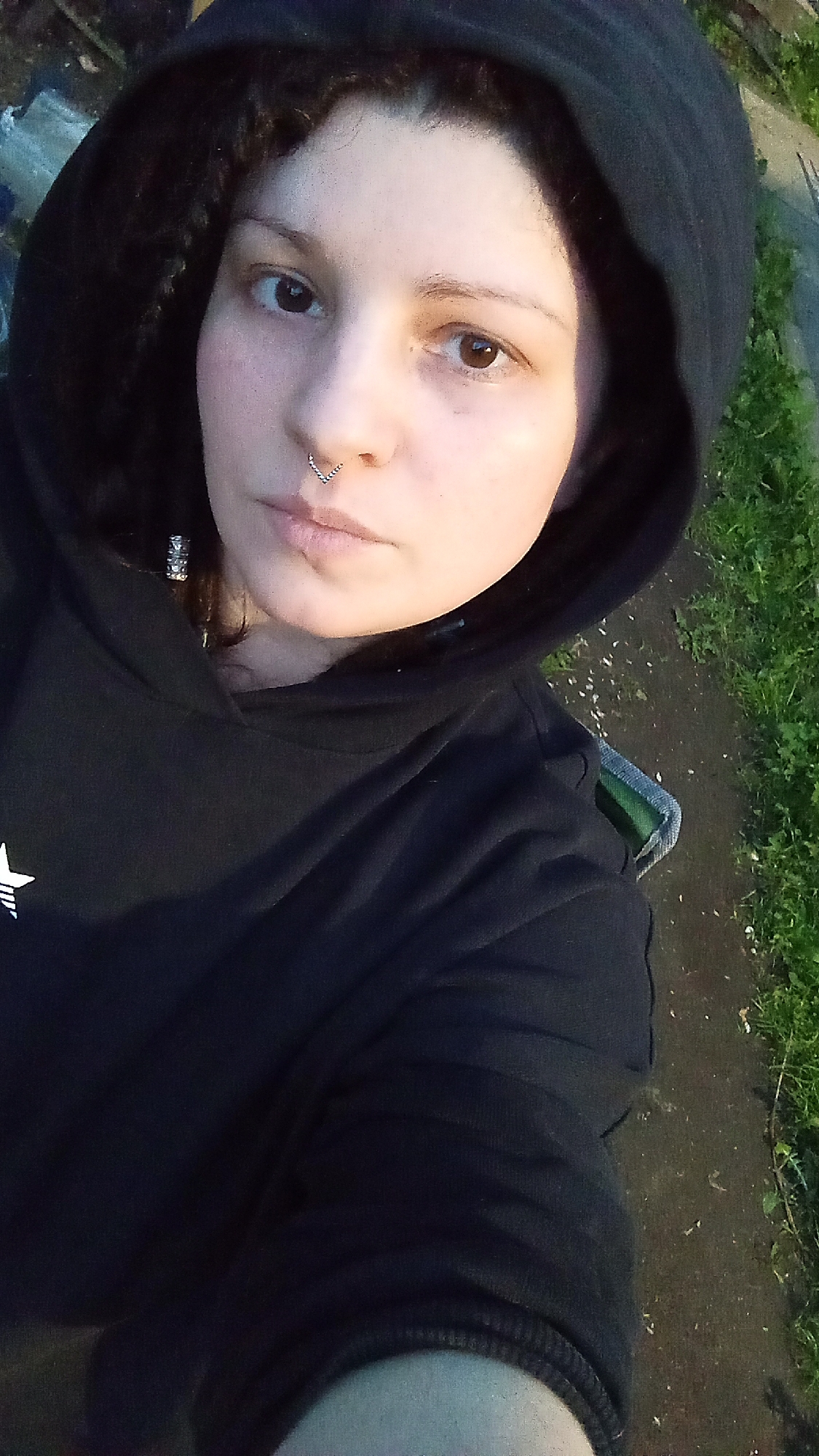A person with light skin, wearing a black hoodie and a silver septum ring, is pictured from a slightly high angle. The person has brown eyes and dark curly hair partially obscured by the hoodie. To the right of the person, there is a paved surface with grass growing along the edge. Based on the metadata, the location is Юдино, Russia.
