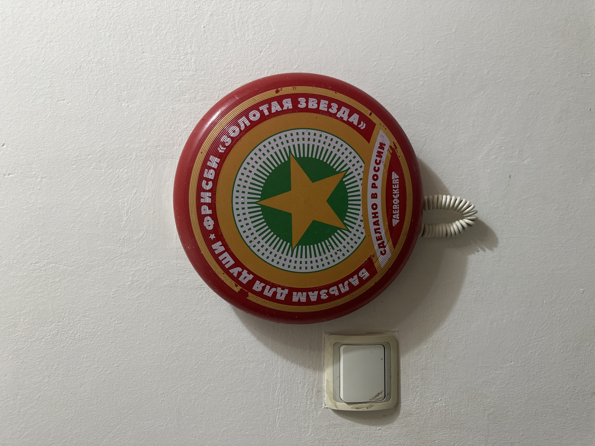 The scene depicts a textured white wall in Budva, Montenegro. Mounted centrally on the wall is a red circular object, approximately 30 cm in diameter, featuring a concentric design. The outermost band is red, followed by a gold band with white Cyrillic text that translates to "BALM FOR THE SOUL * FRISBEE «GOLDEN STAR»." The center features a green circle with a prominent gold five-pointed star. Below the star, additional white Cyrillic text translates to "MADE IN RUSSIA" and "AEROCKER." A white coiled cord extends downwards from the bottom center of this circular object. To the left of the circular object, an off-white electrical wall switch is installed. The switch has a rectangular white button, currently in an unpressed position, set within a slightly discolored off-white bezel. Both the circular object and the switch cast distinct shadows to their right, indicating a light source from the upper left. No individuals or active interactions are present in the image.