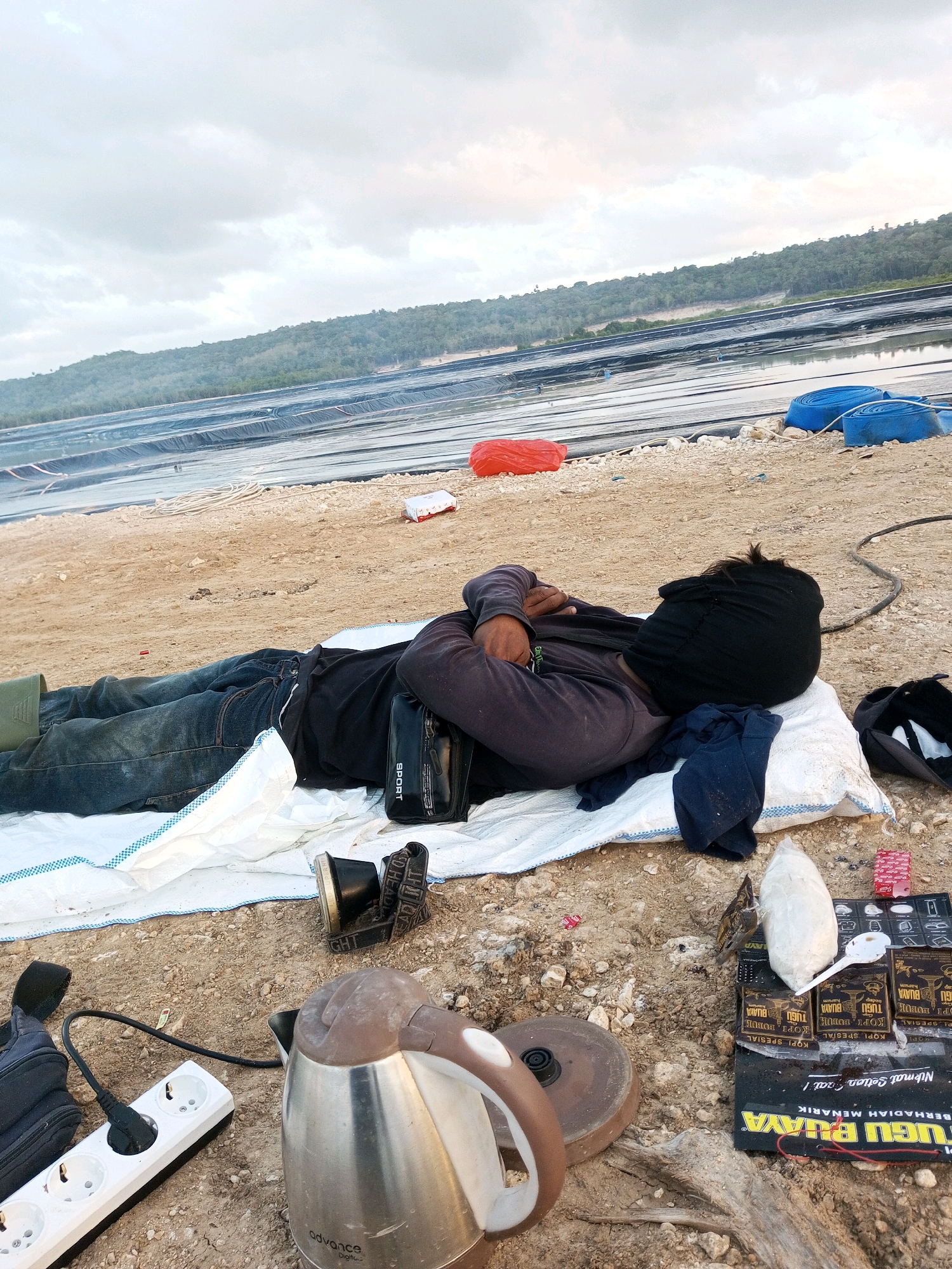 In Matasio, Indonesia, a person lies on their back on a white tarp on a sandy surface. They are wearing a dark long-sleeved shirt, dark pants, and a black head covering that obscures their face. Their arms are crossed over their chest. To the right of the person, a black bag labeled "SPORT" is visible, as is a black baseball cap. In the foreground, a metal electric kettle, a power strip with a plugged-in cable, a black bag with a strap, and a small headlamp are arranged on the ground. Further to the right, several packets of coffee and what appears to be a large white bag of flour or sugar, along with a white spoon, are placed on a black mat. In the background, a body of water is covered with black plastic sheeting, and a tree-covered hill rises beyond it under a cloudy sky. Two large blue coils of hose and a red object are also visible on the ground near the water's edge.