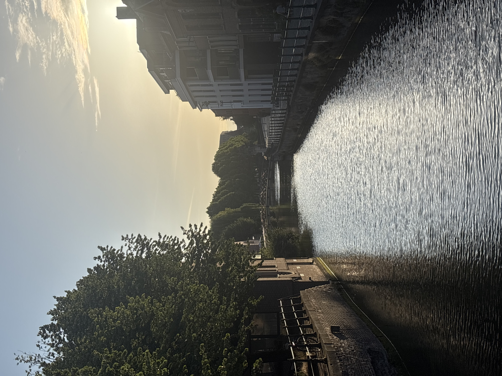 The image depicts a tranquil canal in Ghent, Belgium, at sunset. The sun, low on the horizon, casts a warm glow on the water, creating shimmering reflections. Tall trees line the left bank of the canal, their dense foliage casting shadows. On the right bank, a row of buildings, some with modern architecture and balconies, stands juxtaposed with older brick structures. A narrow walkway with a black railing runs along the canal's edge. A bicycle is parked near a building on the left, and a person is seen walking on the right bank, their back to the camera. In the distance, a bridge spans the canal, and a dense line of trees forms a backdrop. The sky is a pale blue with wispy, golden clouds near the setting sun. The overall impression is one of peaceful urban scenery during the golden hour.