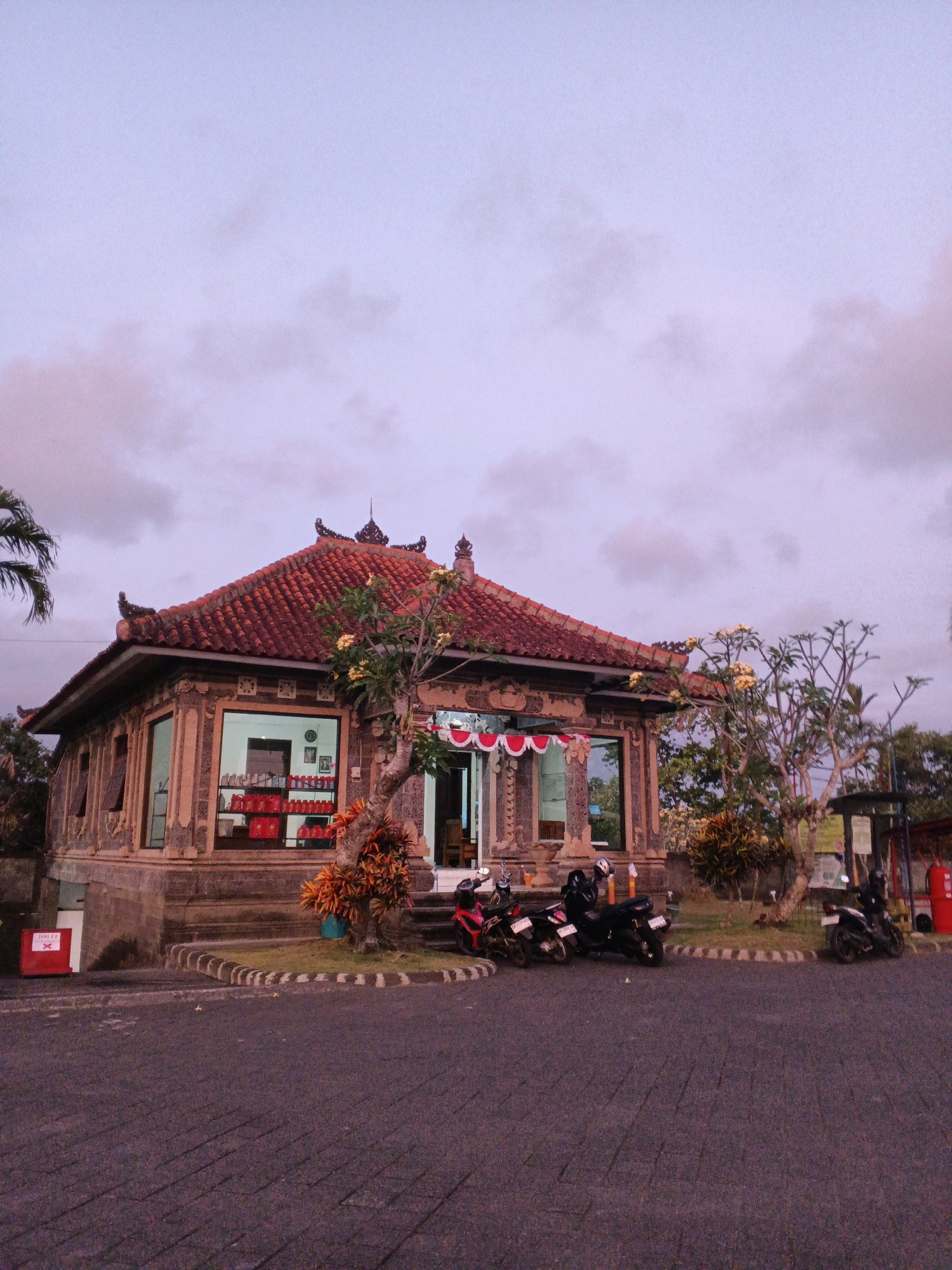 The image depicts a traditional Balinese building with a red tile roof, ornate stonework, and large glass windows. Inside, shelves are stocked with red containers. Several motorcycles are parked in front of the building. Red and white bunting is draped across the entrance. A small tree with yellow flowers is planted to the right of the building, and other vegetation surrounds the structure. The ground in front is paved with dark interlocking stones. A red sign with a white "X" and the word "TOILET" is visible to the left. The sky above is a pale, cloudy purple, suggesting it is either dawn or dusk. The scene appears to be a small shop or refreshment stand, possibly located near a tourist attraction or rest stop in Denpasar, Indonesia. There are no people visible in the image.