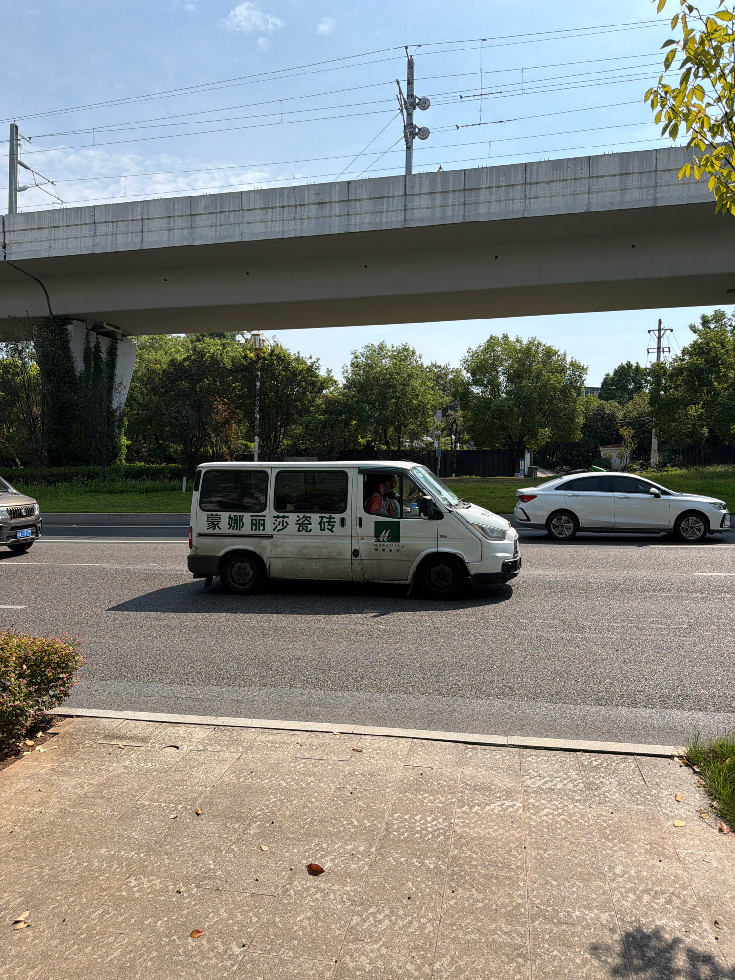 A white van with Chinese characters is driving on a road in Jiangjin, China, beneath a raised overpass that carries train tracks and power lines. The van has the characters "蒙丽莎瓷砖" on its side, which translates to "Mona Lisa Tiles." A white sedan is driving in the lane next to the van, moving in the same direction. The road is asphalt, and a paved sidewalk with scattered leaves is visible to the left of the road. Trees and vegetation line the right side of the road, and the sky is blue with scattered clouds. The overpass is made of concrete and appears to be supported by large pillars.