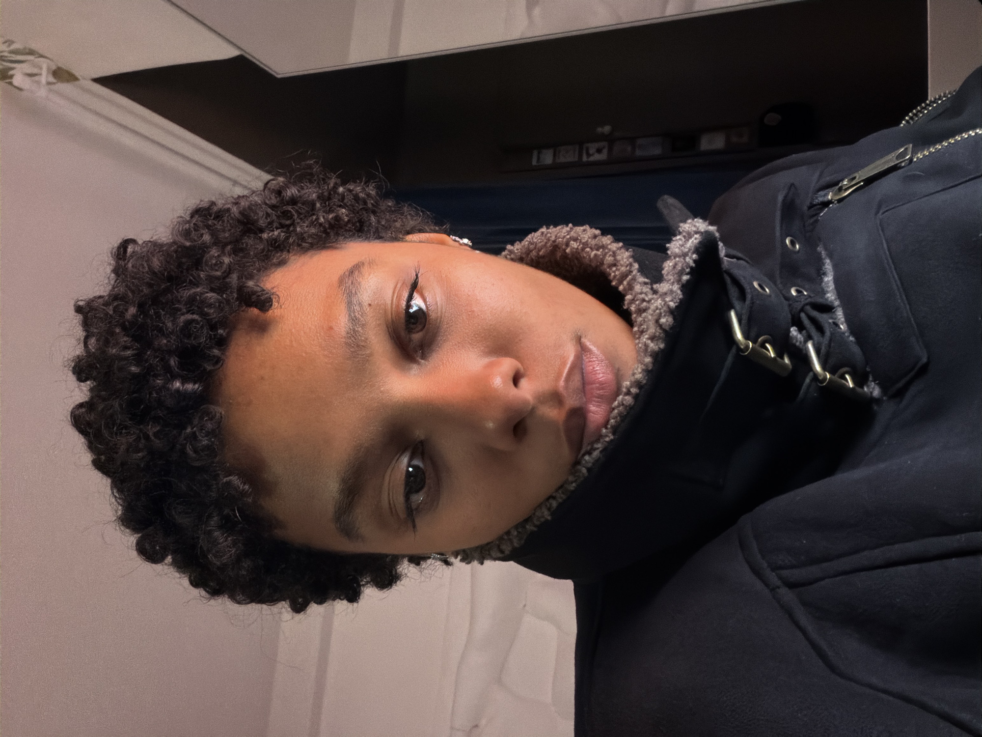 A close-up, slightly rotated image shows a person with dark, curly hair and medium skin tone. They are wearing a dark jacket with a shearling-lined collar and metal hardware. The background consists of a light-colored wall and a darker, undefined space. The setting does not obviously indicate Paris, France, or any other specific location.