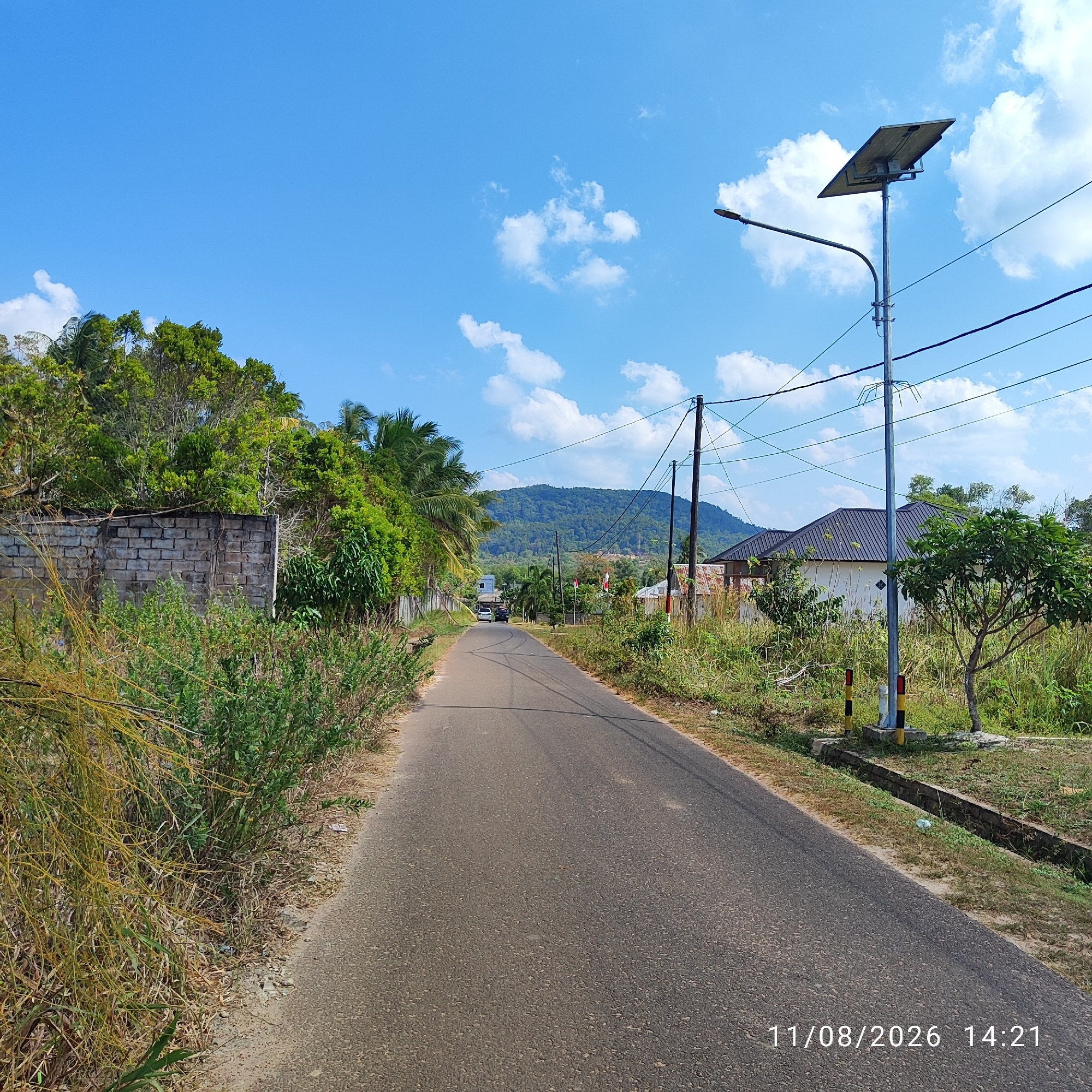 An eye-level, medium shot captures a paved road in the city of Pembaharuan, Indonesia, on a bright, clear day. The road, which runs into the distance, is flanked by lush green vegetation and a grey brick wall on the left, and a grassy area with a modern house and a solar-powered street light on the right. In the background, a verdant, tree-covered hill dominates the horizon under a brilliant blue sky dotted with scattered white clouds. A few cars are visible on the road, and the date stamp "11/08/2026 14:21" is overlaid in the bottom right corner of the image.