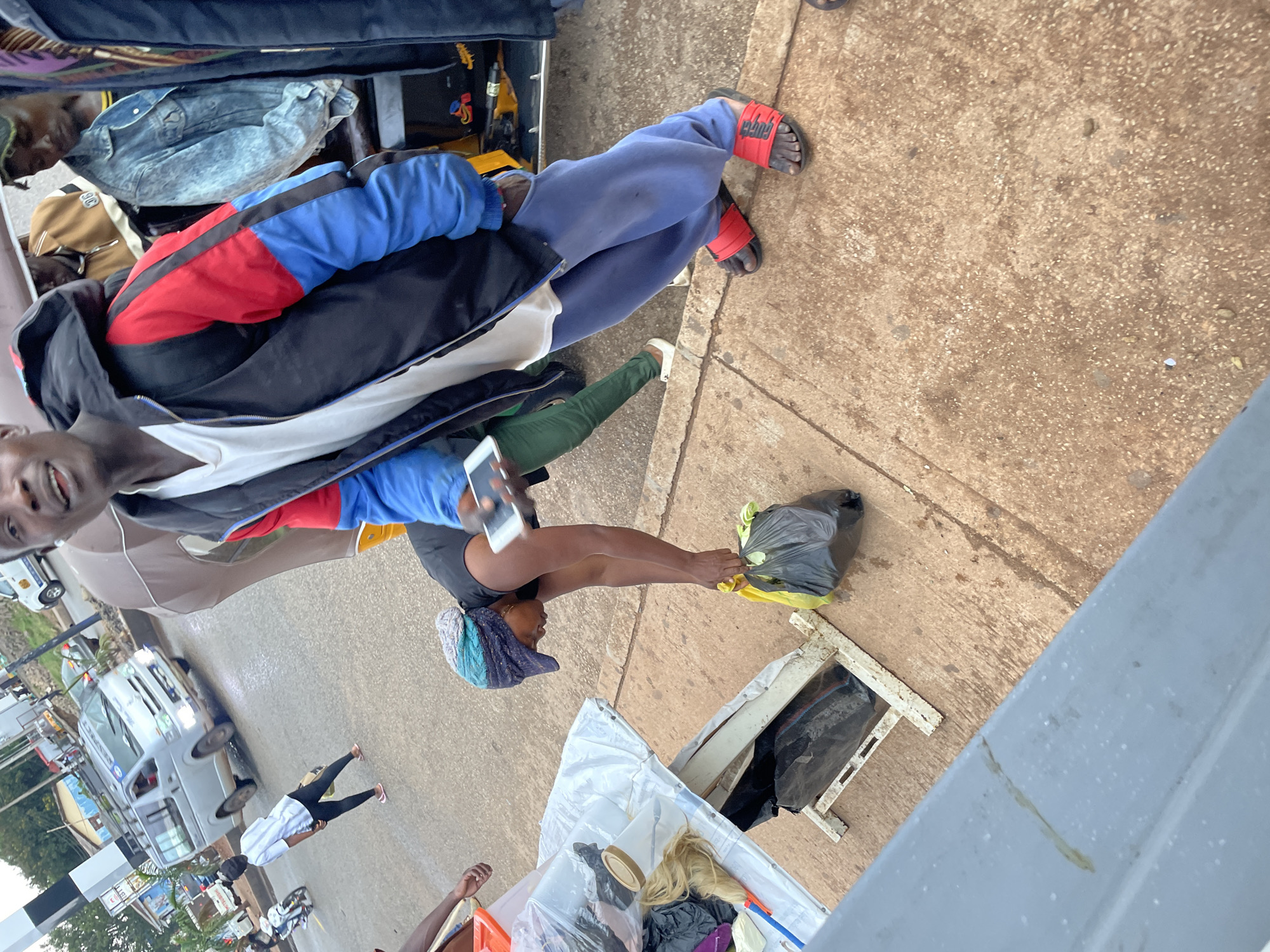 This is a street-level, eye-level shot taken on a cloudy day in Jos, Nigeria. In the foreground, a man wearing a blue and red jacket and blue sweatpants is looking at the camera and holding a smartphone. He is wearing red sandals. To his left, a woman wearing a headwrap and a black t-shirt is bending down to pick up a black plastic bag filled with items. Next to her is a white table with various items displayed, including plastic bags, a wig, and a container. In the background, a silver van with "RESPECT" written on it is driving on the street, along with a few motorcycles and other people. The environment is an urban setting with a paved road and sidewalk.