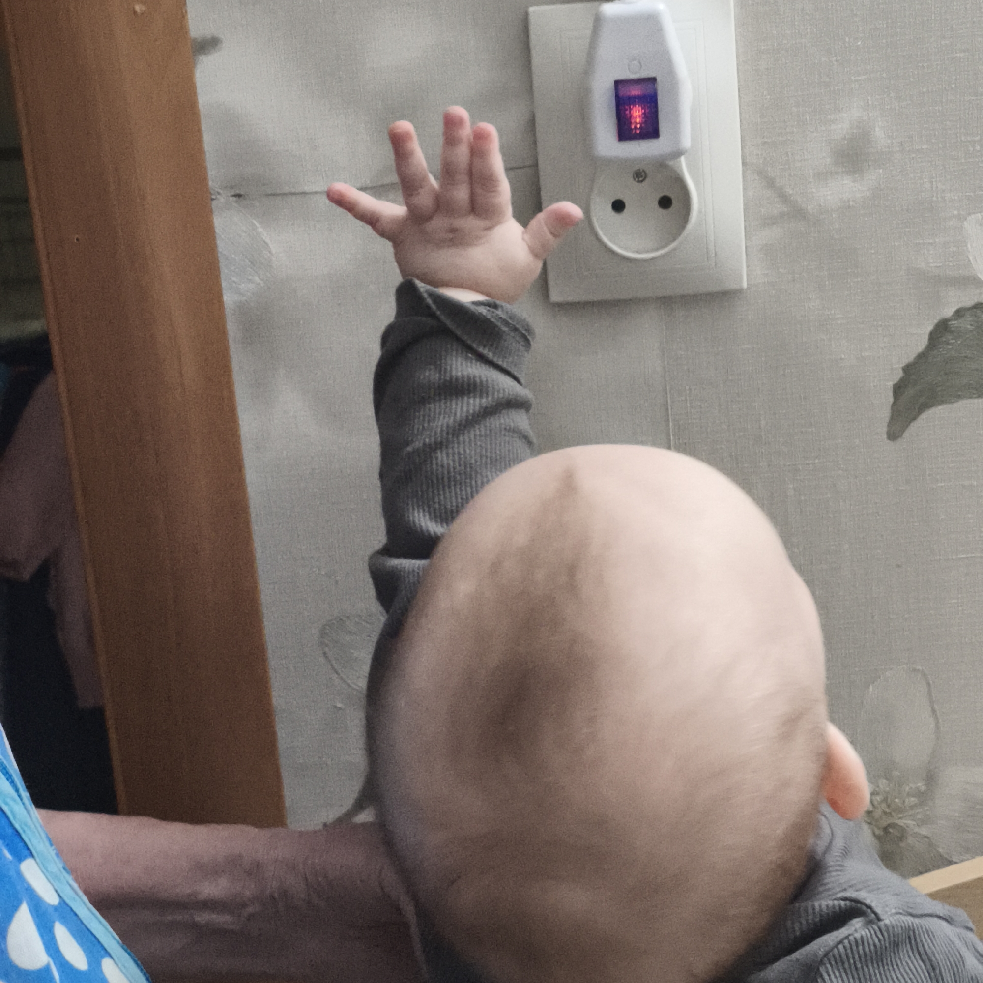 A small child reaches towards an electrical outlet with a night light plugged into it. The child has a bald head and is wearing a gray, long-sleeved shirt. A hand and arm of an adult are visible in the bottom left corner, possibly holding or supporting the child. The outlet is mounted on a wall with a floral pattern. The photo was taken indoors. The city of Chaikovsky, Russia is provided as the location.