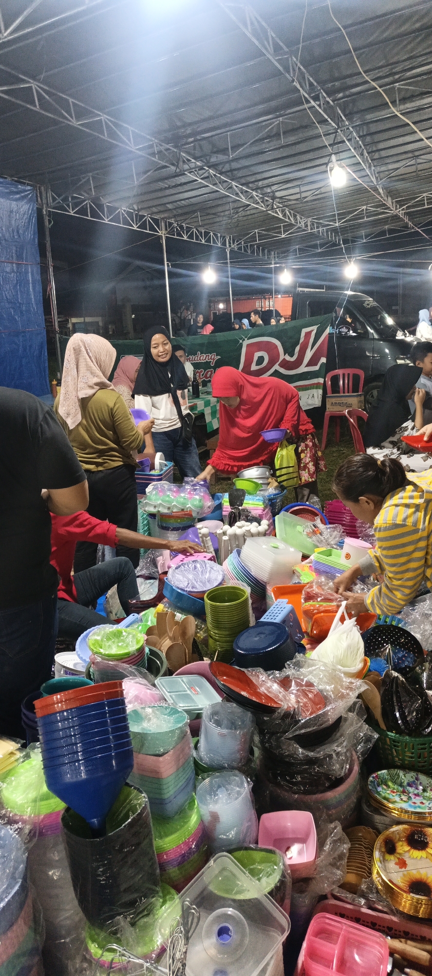This image depicts an outdoor market stall in Banding Agung, Indonesia. A variety of plastic household items, primarily bowls and containers in various colors, are displayed for sale. Several people, some wearing headscarves, are present at the stall, appearing to be customers and vendors. The stall is covered by a large, tent-like structure supported by metal beams. In the background, a vehicle is visible, along with more market stalls and structures. The lighting suggests it is either evening or nighttime. The overall scene captures a bustling marketplace atmosphere.