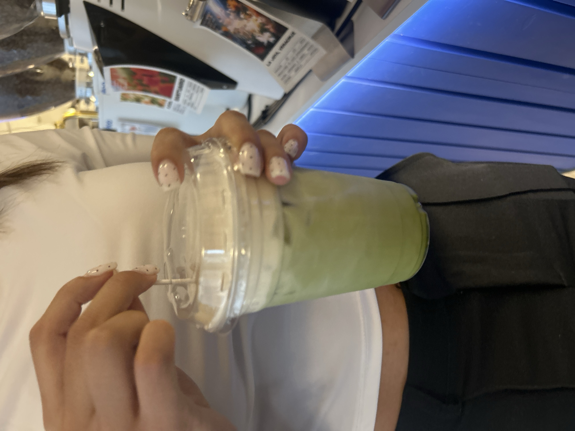 A person in a white top stirs a green beverage with ice and a lid in a clear plastic cup, using a straw. The person has manicured fingernails painted with white polish and small pink polka dots. The person is wearing black pants. A piece of restaurant equipment is partially visible in the background, as well as a blue-colored overhead structure. I cannot definitively state that this image was taken in San Bernardino Tlaxcalancingo, Mexico, as there is no specific evidence to support that.
