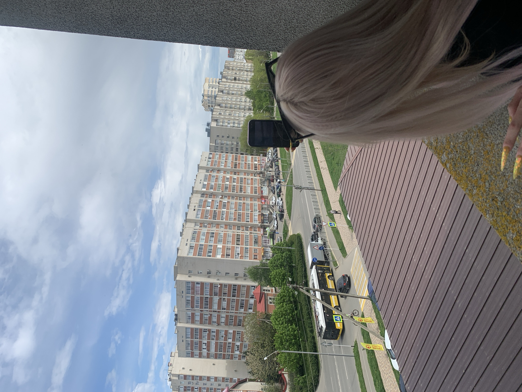 A woman with blonde hair is standing on a brown balcony, holding a phone in front of her face. She is wearing glasses and has long, yellow fingernails. In the background is a city street with a tall apartment building, a yellow bus, and several parked cars. The sky is partly cloudy.