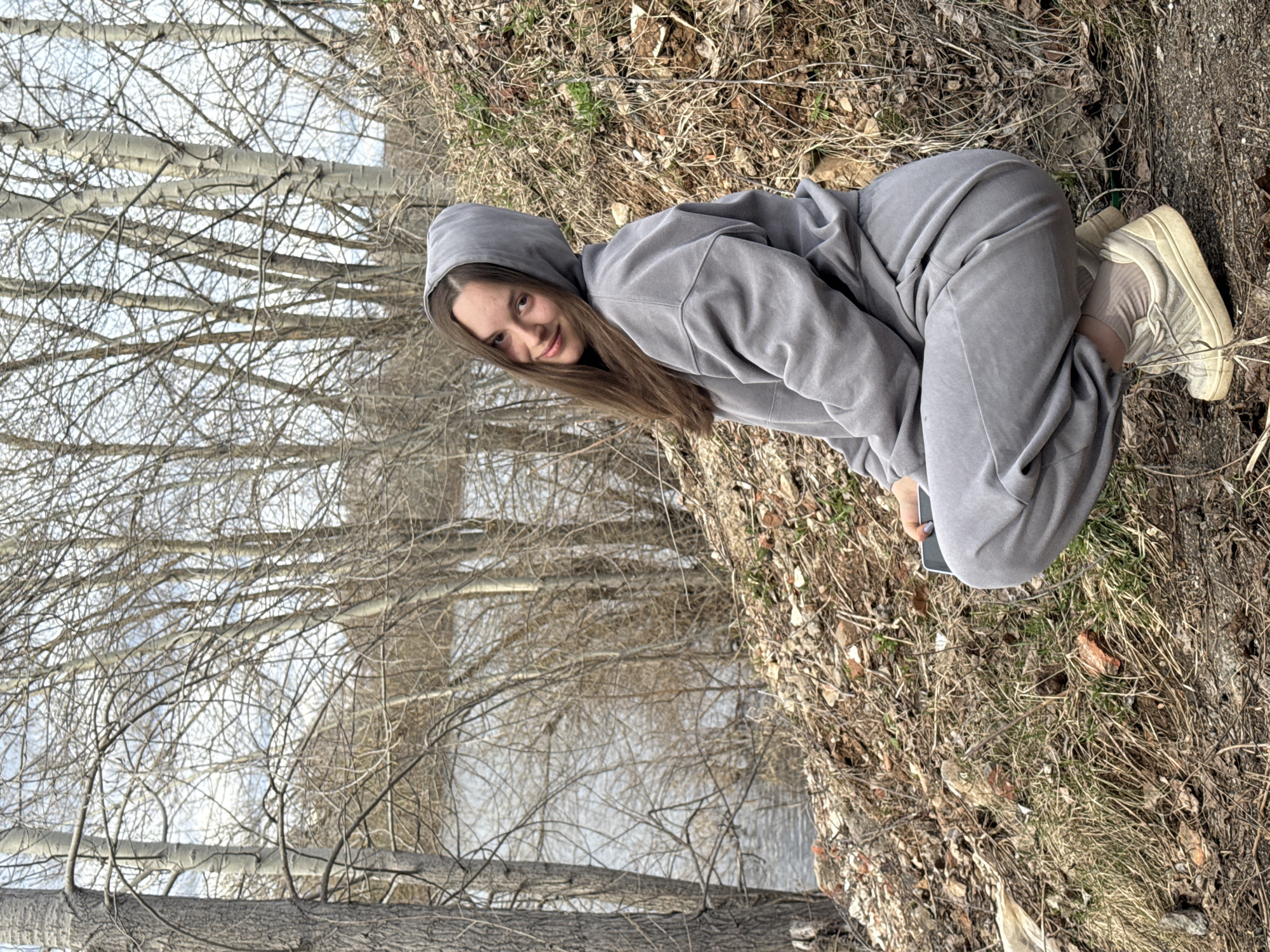 A young woman with long brown hair is crouching on the bank of a river in what appears to be a natural, undeveloped area. She is wearing a gray hooded tracksuit and light-colored sneakers. Her head is tilted slightly, and she is looking directly at the camera with a slight smile. She is holding a dark-colored object, likely a phone, in her hand. The ground is covered in dry leaves, grass, and twigs. In the background, there are bare trees and a body of water, suggesting a cold or winter season. The location is Chaikovsky, Russia, as the description specifies.