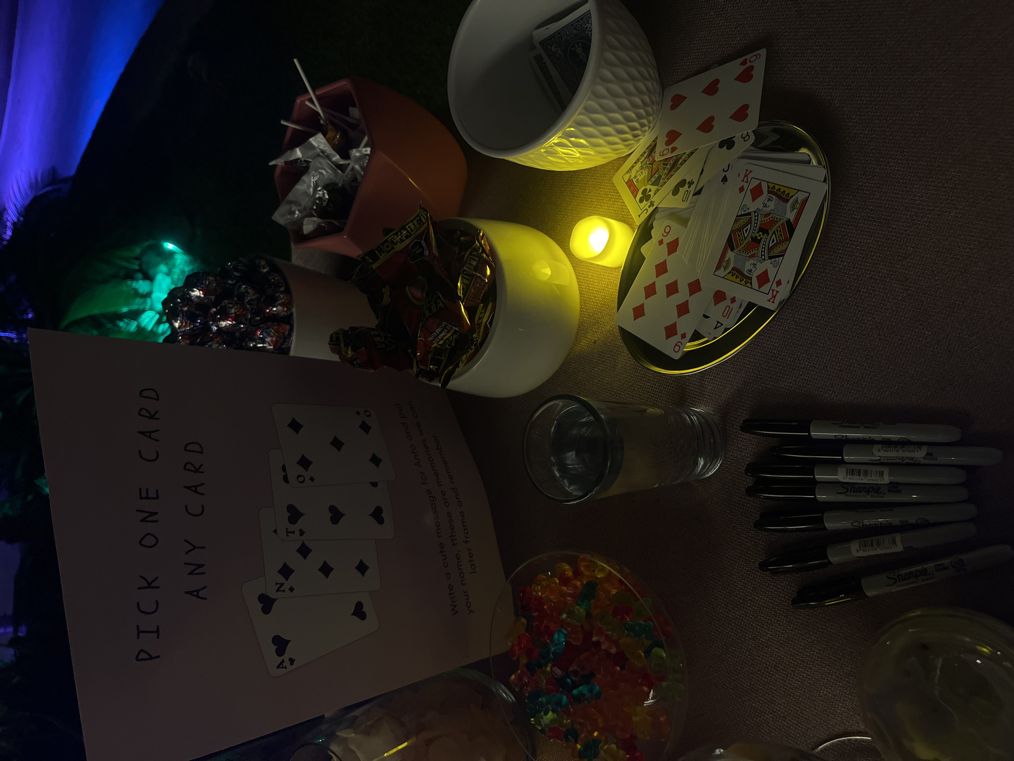 The image shows a tabletop display with various objects intended for a party or gathering. There is a sign encouraging attendees to pick a playing card and write a message on it for someone named "Anto." Beside the sign, there are playing cards, a small tea light candle, a glass of water, a collection of black Sharpie markers, gummy bears in a glass bowl, and two different containers filled with candy. Other containers filled with other treats are also present. The location is suggested to be San Bernardino Tlaxcalancingo, Mexico, but there is no visual information confirming this place in the picture.