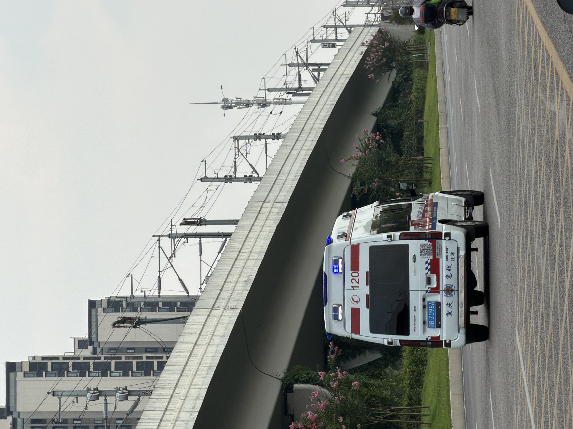 An ambulance is driving on a road in Jiangjin, China, under an elevated highway. The ambulance is white with red and blue markings. The license plate reads "B-Z098A". On the back of the ambulance, there is a red circle with "120" inside it, which is the emergency number in China. There are also Chinese characters that say "重庆" (Chongqing) and "急救" (Emergency). To the right of the ambulance, a person on a motorcycle is also driving on the road. In the background, there are tall buildings and power lines. The sky is overcast.