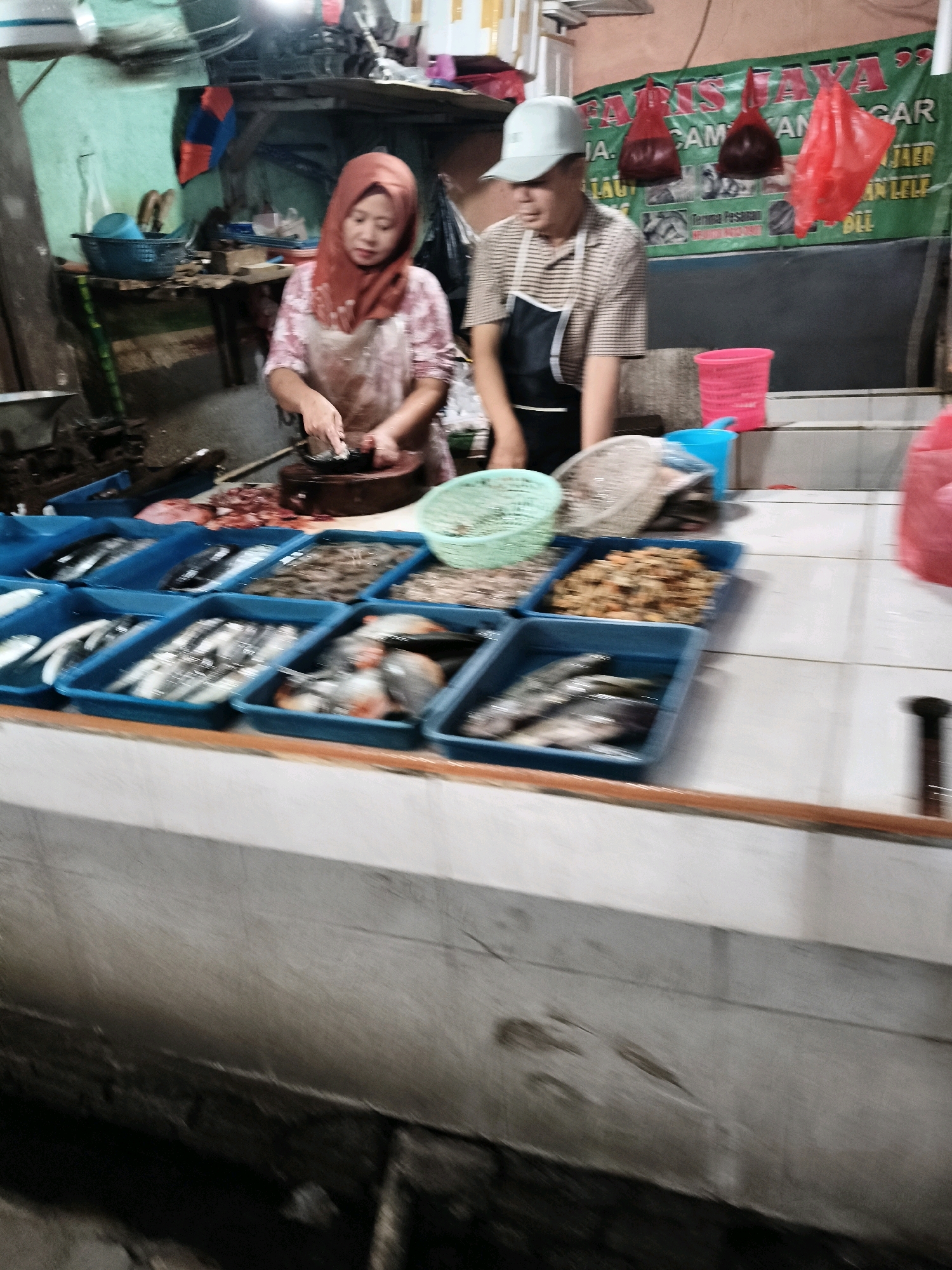 The image depicts a market scene in Setiamekar, Indonesia, focusing on a stall where seafood is being prepared and displayed. Two individuals are visible behind a counter. A woman, wearing a red hijab and an apron over a patterned shirt, is actively engaged in preparing food, her hands resting on a chopping board. To her right, a person in a striped shirt and a light-colored cap is also behind the counter, appearing to be involved in the same activity.
The counter displays a variety of fresh seafood, predominantly fish and shrimp, arranged in blue plastic containers. Some containers hold whole fish, while others contain processed seafood or shrimp. There are also green and white woven baskets on the counter, one of which contains shrimp. To the left of the scene, a dark-colored weighing scale is partially visible.
In the background, a green banner with red lettering hangs, likely advertising the business. Red plastic bags are suspended from above, and other market stalls or structures are suggested by the shelves and wall in the background. The overall environment appears to be an indoor market with a focus on seafood sales.