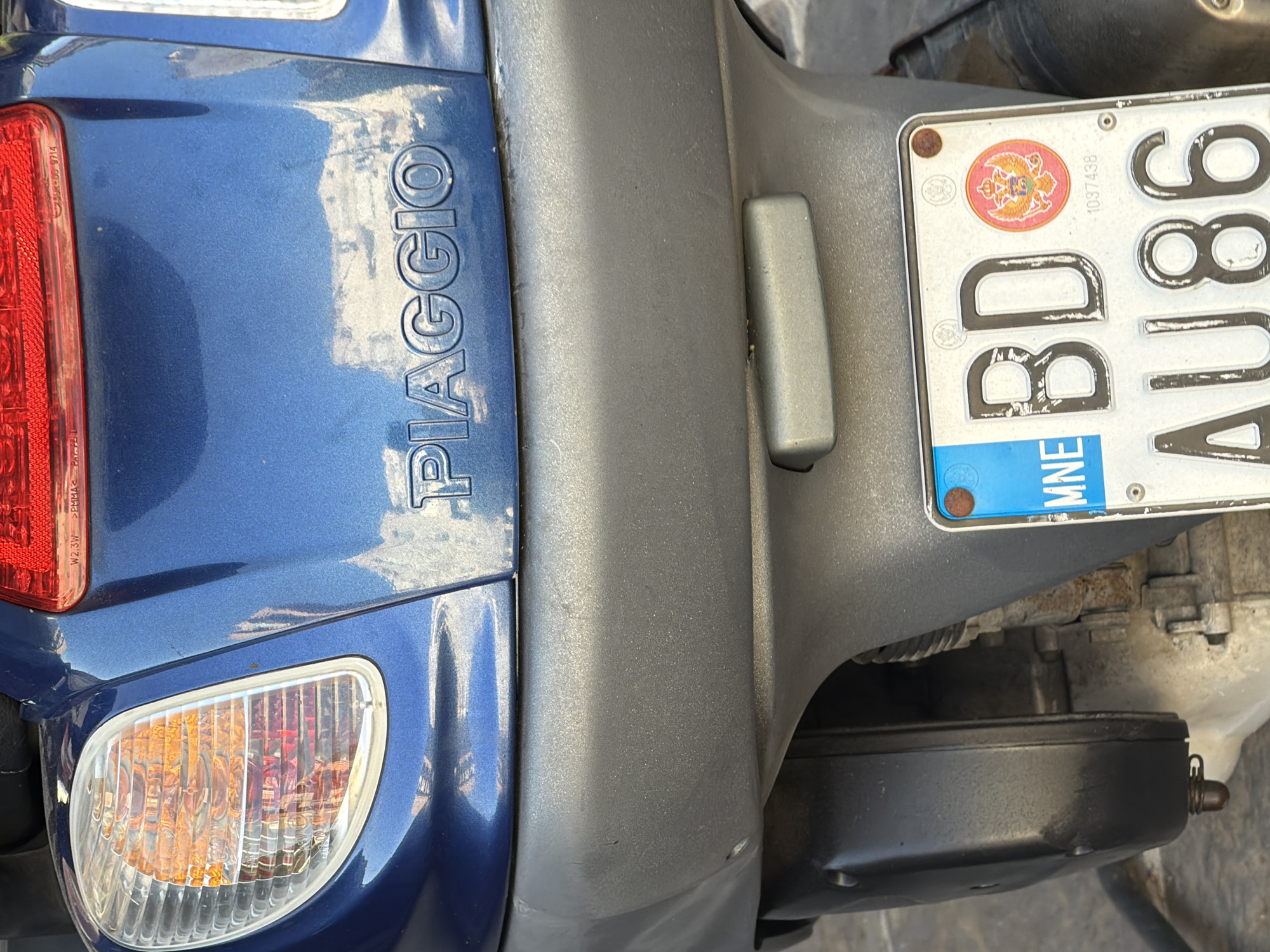 The image displays the rear section of a blue Piaggio scooter parked in the city of Budva, Montenegro. Prominently featured is the rear license plate with the characters "BD AU86" and a Montenegrin coat of arms. The scooter's taillight and turn signals are visible, along with parts of the engine and a side stand. The background suggests an outdoor urban setting, with a weathered ground surface. The scooter is the sole subject of the image.