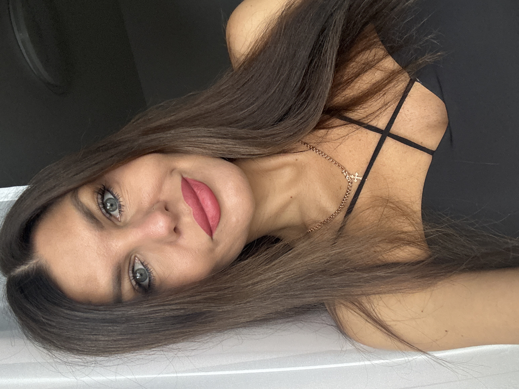 A close-up selfie shows a woman with long, dark brown hair and light blue eyes. She is wearing a black top with a criss-cross neckline and a rose gold chain necklace with a cross pendant. Her makeup includes pink lipstick and mascara. The background is out of focus, with a white curtain on the left and a dark surface on the right. The image appears to be taken indoors. There is no discernible information to confirm the location in Chaikovsky, Russia.
