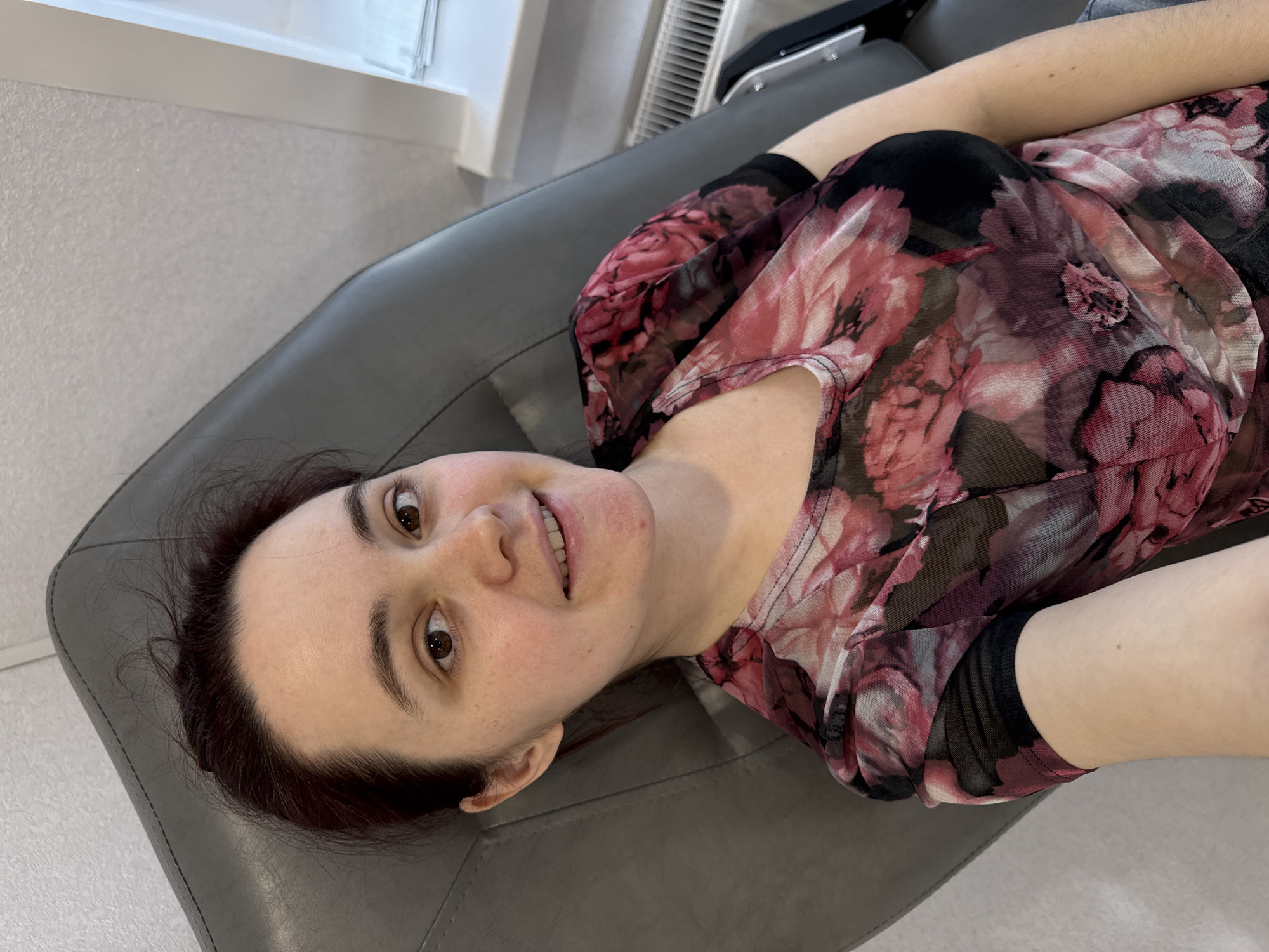 A woman with dark hair and fair skin is lying on a gray leather couch. She is wearing a sheer blouse with a floral pattern in shades of red, pink, and black. The location is indeterminate and there is no information available in the image to establish the city of Izhevsk, Russia as the location. Other objects in the background include a radiator and a window.