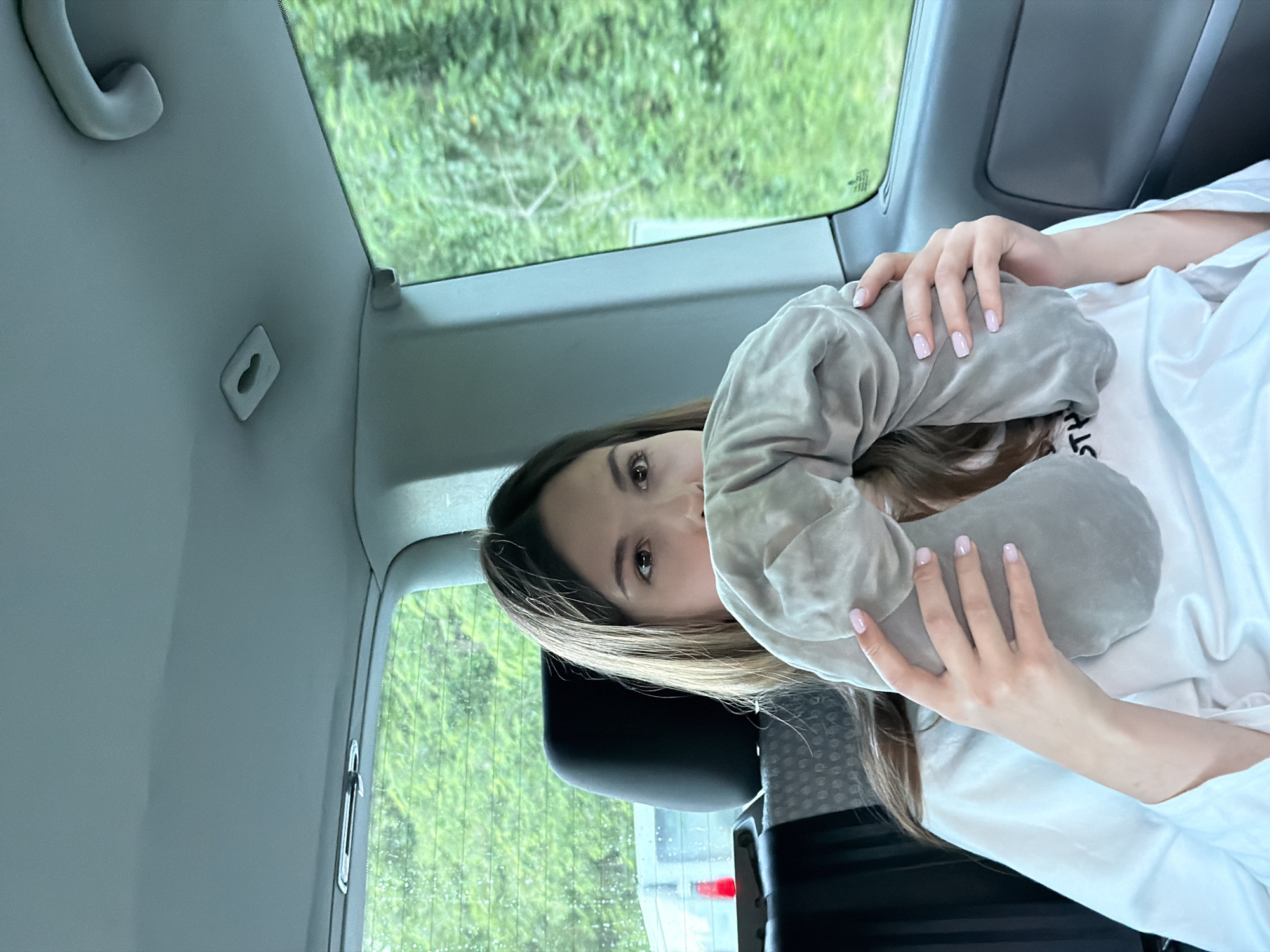 A female passenger sits in a car, holding a grey travel neck pillow, possibly preparing for a trip. The background shows greenery through the car window. This image was taken in Mali Zvornik Municipality, Serbia.