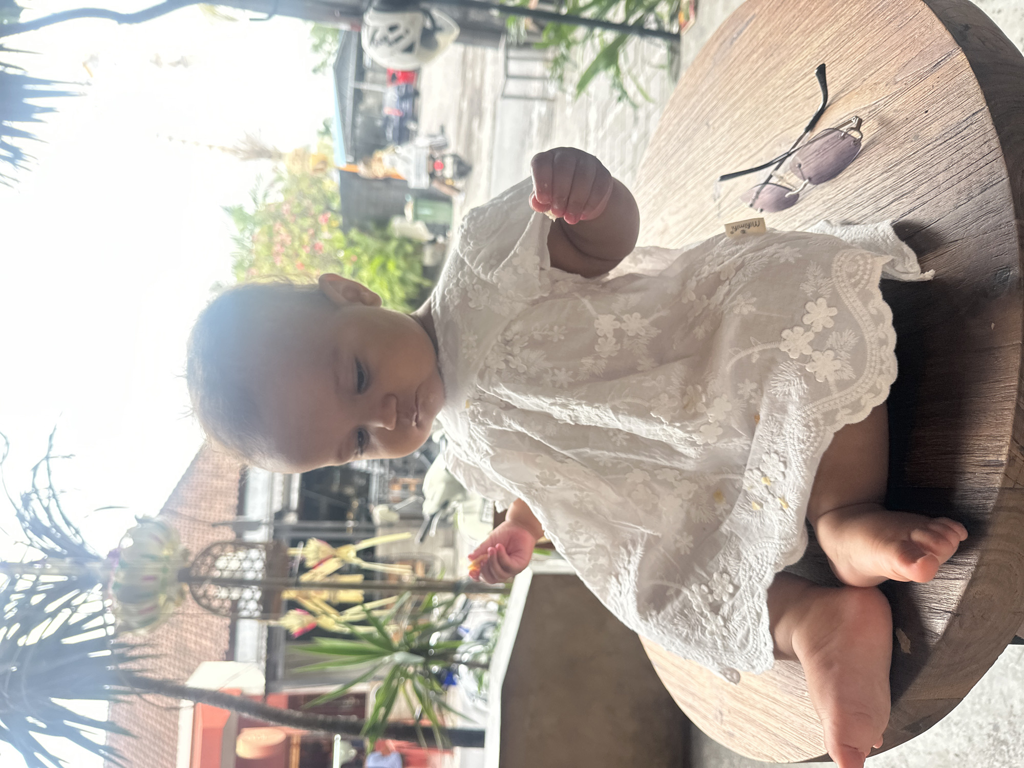 A baby wearing a white lace dress is sitting on a wooden table outdoors. The baby is looking down at a piece of food in its hand. In the background, there are palm trees, buildings, and people. The scene appears to be in a tropical location, possibly a cafe or restaurant in North Kuta, Indonesia. Sunglasses are lying on the table next to the baby.