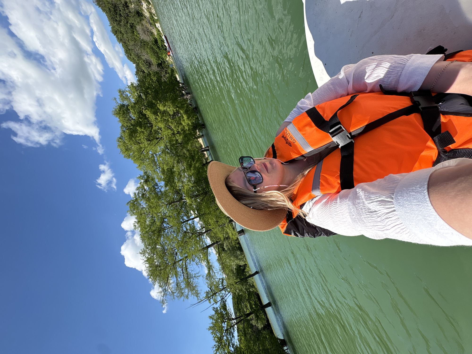 This media file captures a bright outdoor scene in Sukko, Russia, featuring a person relaxing on a body of water. The image is taken from a slightly tilted perspective, showing a vibrant landscape under a clear sky.
In the foreground and to the right, a person is seen from a slightly elevated angle, leaning back on what appears to be a white boat or raft. They are dressed in a white, textured long-sleeved top, an orange life vest, a straw hat, and dark sunglasses. The life vest prominently displays the text "OMUT" and a small fish logo. The person's expression appears relaxed and peaceful.
The central and left parts of the image are dominated by calm, greenish water, exhibiting gentle ripples across its surface. Along the water's edge, lush green trees, possibly cypress varieties, form a dense natural barrier, with some trunks and branches extending into or near the water.
Above the treeline, a vast expanse of clear blue sky is visible, dotted with several bright white cumulus clouds. The lighting suggests it is bright daylight, likely mid-morning or afternoon, contributing to the overall serene and pleasant atmosphere. The scene depicts a leisurely activity on the water, enjoying the sunny weather and natural surroundings.