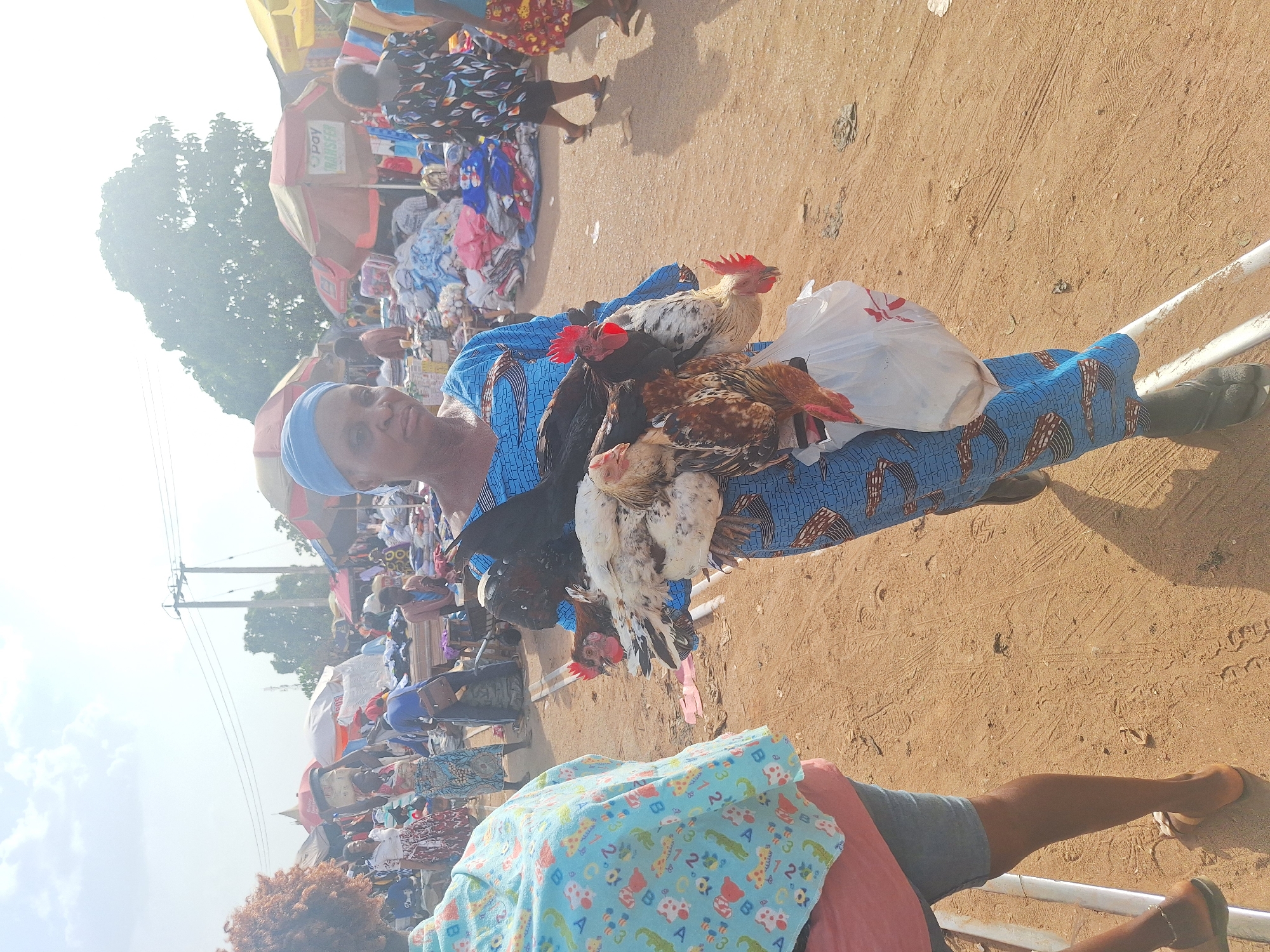 In an outdoor setting at a likely market in Kakuri Gwari, Nigeria, a woman wearing a blue patterned dress and head covering carries several live chickens. The chickens are a mix of colors - white, black, and brown. She also carries a white plastic shopping bag. The area around her appears to be sandy or dusty. In the background, there are numerous people and market stalls with various goods displayed under awnings. Power lines are visible in the sky. Another person, wearing a blue patterned shirt, grey shorts and pink shirt, is partially visible in the foreground.