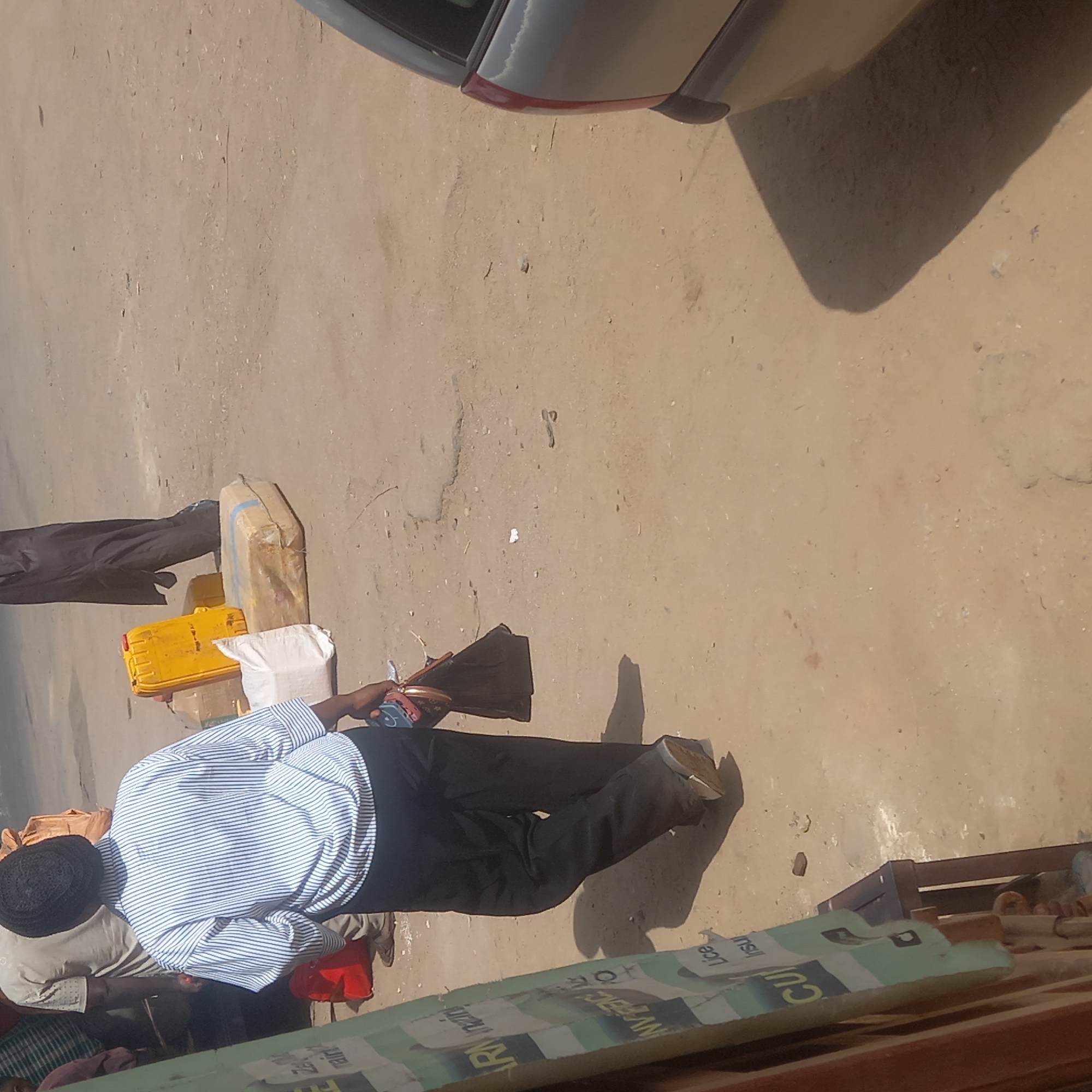 The image, taken in Dadi Riba, Nigeria, captures an outdoor scene under bright daylight. The ground is a light, dusty brown, characteristic of unpaved or lightly paved surfaces. Strong shadows indicate a sunny day, likely mid-morning or afternoon.
In the upper right portion of the image, the rear quarter panel and taillight of a dark-colored car are visible. A prominent shadow from the car extends onto the ground.
Towards the center and left, several objects are placed on the ground next to what appears to be another person's legs (dressed in black trousers). These objects include a large, tan-colored package, possibly a sack, wrapped in what looks like brown paper or fabric with a blue stripe. Next to it is a bright yellow jerrycan, and a white sack or bag. Another black, folded item, possibly fabric or a bag, lies nearby.
The main subject is a person standing, viewed from behind and slightly to the left. This individual wears a blue and white striped long-sleeved shirt, black trousers, and what appears to be a dark, possibly black or navy, cap or kufi. They are holding a brown and pink patterned purse or bag in their right hand, with a mobile phone visible in the other hand. Their shoes are light-colored, possibly worn, with distinct shadows cast by their legs.
In the bottom foreground, a green and white banner or sign is partially visible. Fragments of text can be deciphered, including "CU," "insu," and "Lice," suggesting it might be an advertisement or informational sign. Another person's leg, possibly wearing a red garment, is partially visible below the main subject.
The overall scene suggests a casual street or market setting where people are either waiting, loading, or unloading goods, possibly related to transportation or commerce.