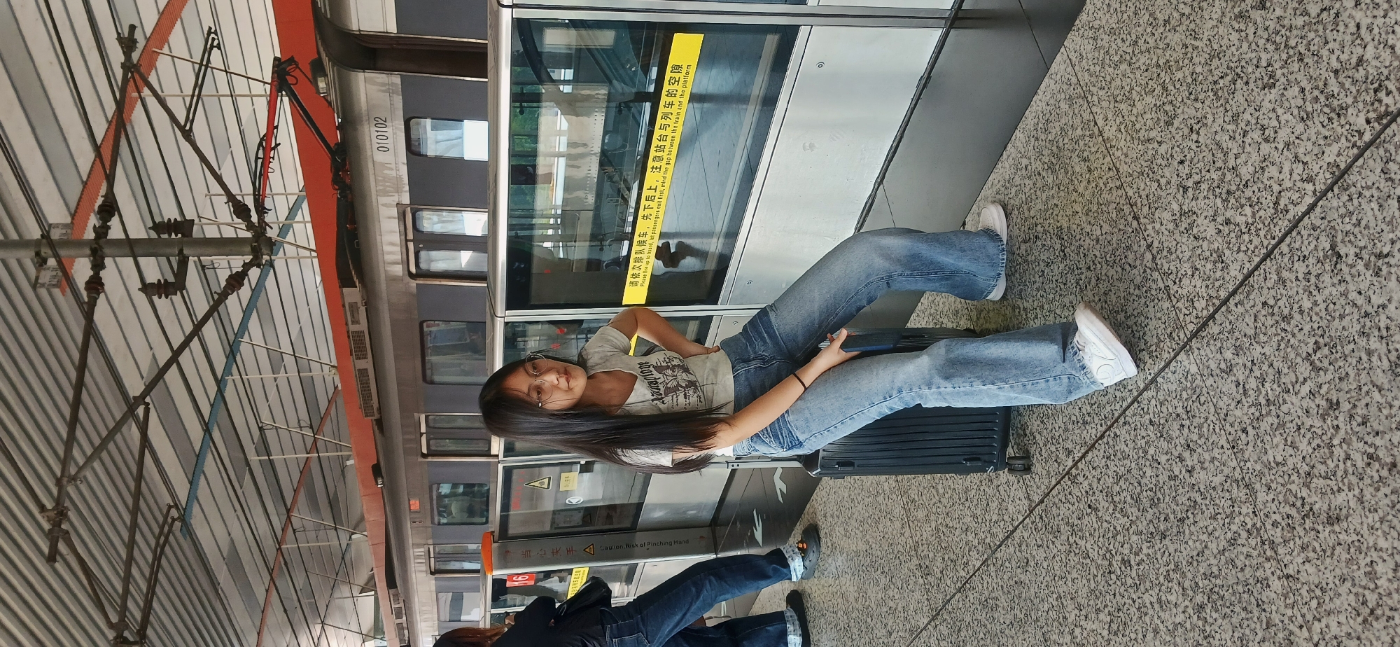 The image captures a casual moment on a train station platform, specifically identified as Chongqing High-tech Zone, China. The primary subject is a young woman with long, dark hair and glasses, seated comfortably on a black hard-shell suitcase. She is dressed in a light-colored t-shirt with a graphic design, blue flared jeans, and white sneakers, and she is looking directly at the camera with a slight smile, appearing relaxed.
The setting shows a train waiting alongside the platform, partially visible through the transparent platform screen doors. The train is dark grey with a lighter stripe and has the number "010102" displayed on its side. The platform floor is made of light grey speckled tiles with dark grout lines. Above, intricate metallic structures, likely part of the station's roof or catenary system, are visible against a light background. Another person, seen from the back, is partially visible at the bottom left, also appearing to be seated and waiting.
Several pieces of text are visible on the platform screen doors. A prominent yellow strip displays both Chinese and English text, instructing passengers: "请依次排队候车,先下后上,注意站台与列车的空隙 Please line up to board, let passengers exit first, mind the gap between the train and the platform." Another sign, yellow and black, warns of potential danger with "当心夹手 Caution. Risk of Pinching Hand." The scene conveys a typical wait at a modern urban transit station during what appears to be daytime.