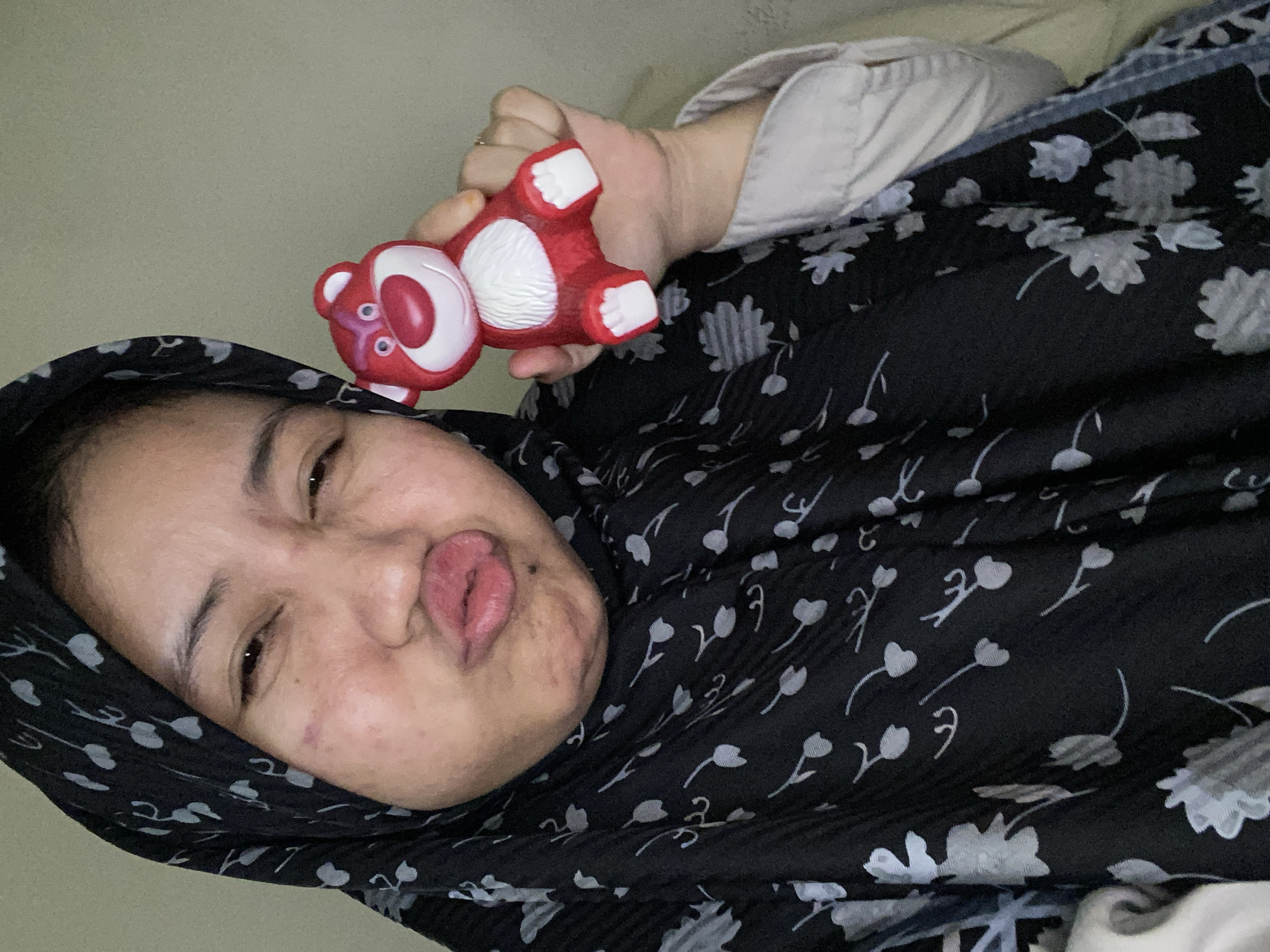 A young woman wearing a black floral hijab and a light-colored button-down shirt is holding a red Lotso bear toy from Toy Story. She is making a kissing face at the camera and her eyes are squinting. The background is a plain, light-colored wall. The image appears to be a selfie taken indoors, likely in a residential setting in Banjarbaru, Indonesia.