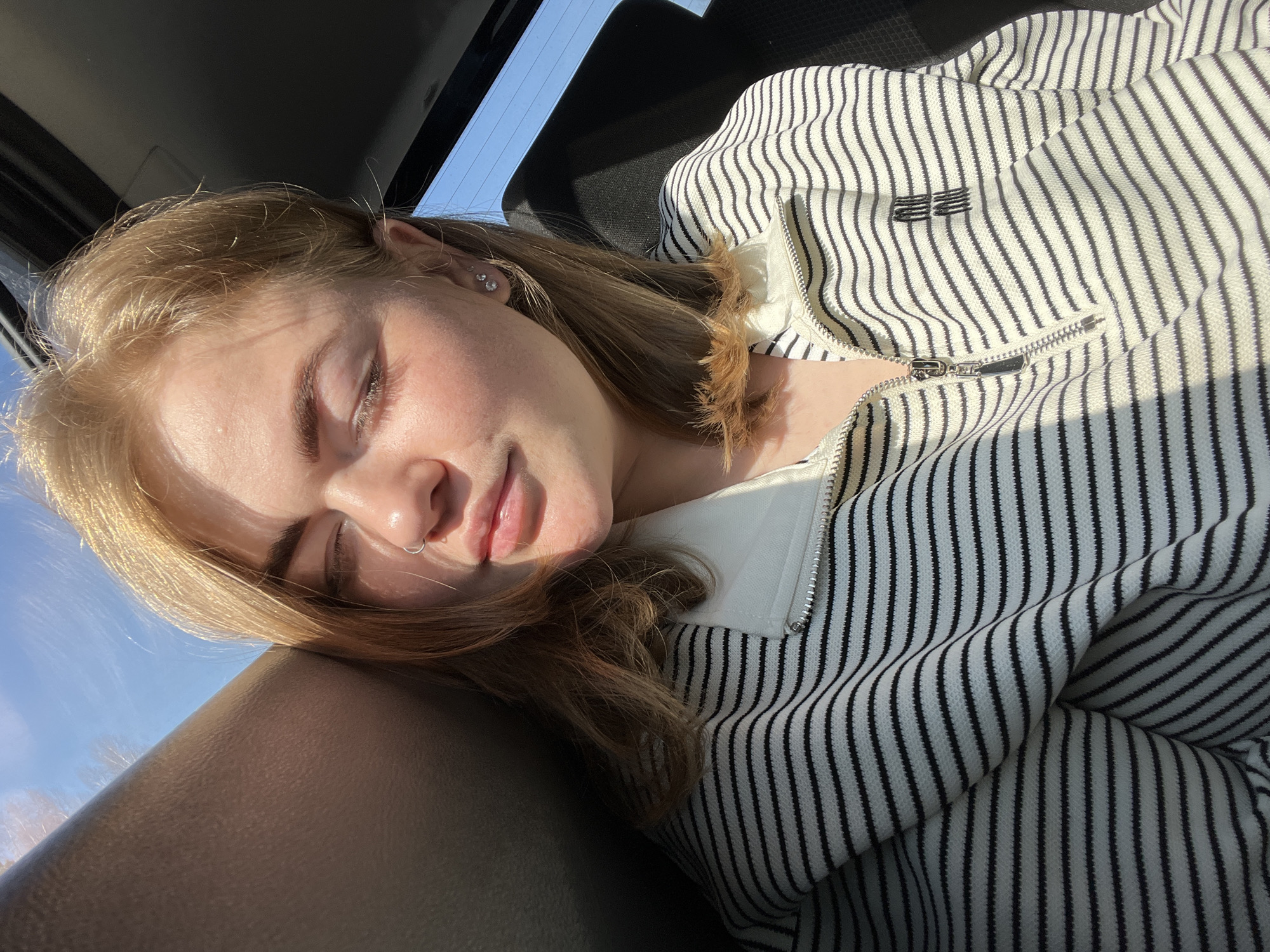 A young woman is reclining in what appears to be a vehicle, possibly a car, as suggested by the headrest and a portion of a window visible in the background. She is lying on her side, head resting on the headrest, eyes closed or partially closed. She has blonde hair and a nose ring. She is wearing a striped black and white zippered top. The image does not contain enough information to determine the city of Krasnokamsk. The scene is well-lit, with sunlight falling on the woman's face and clothing.