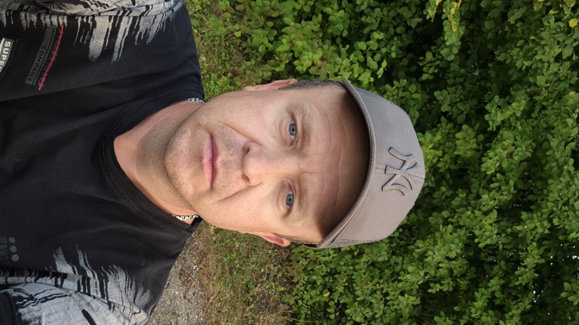 In Ergolding, Germany, a man in his late 40s or early 50s is photographed from the chest up. He has fair skin, light blue eyes, and short, light brown hair. He is wearing a gray baseball cap with the "NY" logo and a black t-shirt with a gray and black abstract pattern on the sleeves. A silver chain is visible around his neck. The man is facing the camera with a neutral expression. He is standing outdoors with a backdrop of lush green foliage and some dry ground visible in the lower right corner.