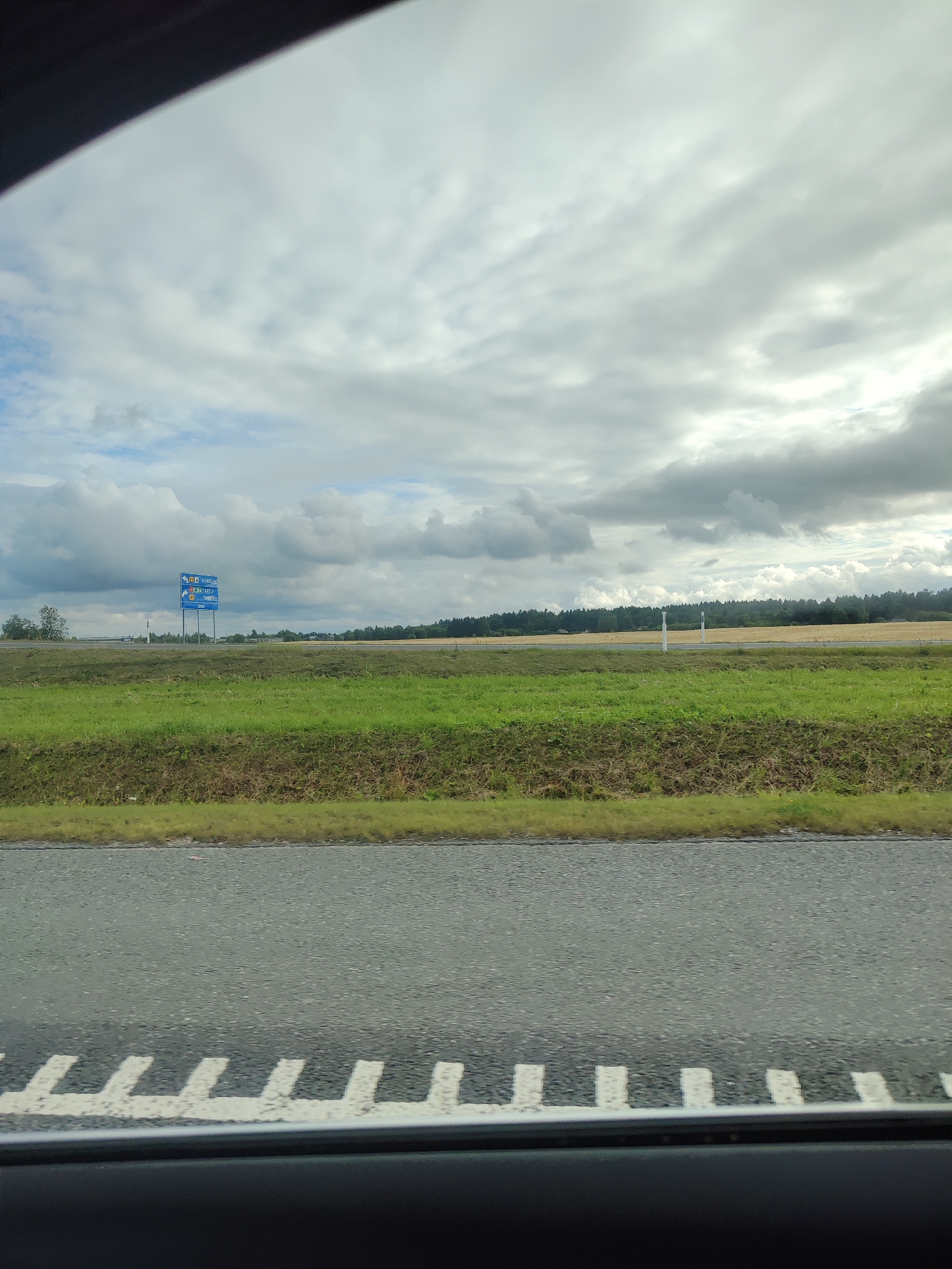 A roadside view captures a landscape under a cloudy sky. In the distance, a blue directional signpost with white text and symbols stands prominently, indicating routes to "Täkumetsa," "Kallaste," and "Tartu." The sign also displays route numbers "7" and "39". Adjacent to the sign, a sparse cluster of trees and a few buildings are visible, nestled against a backdrop of a dark green forest bordering a field of dry, yellowing crops. In the foreground, a green grassy embankment runs along the edge of a grey asphalt road, marked with white dashed lines indicating a lane division. The sky is filled with a mix of grey and white clouds, suggesting overcast weather. The image is taken from a vehicle, as indicated by the dark frame at the bottom.