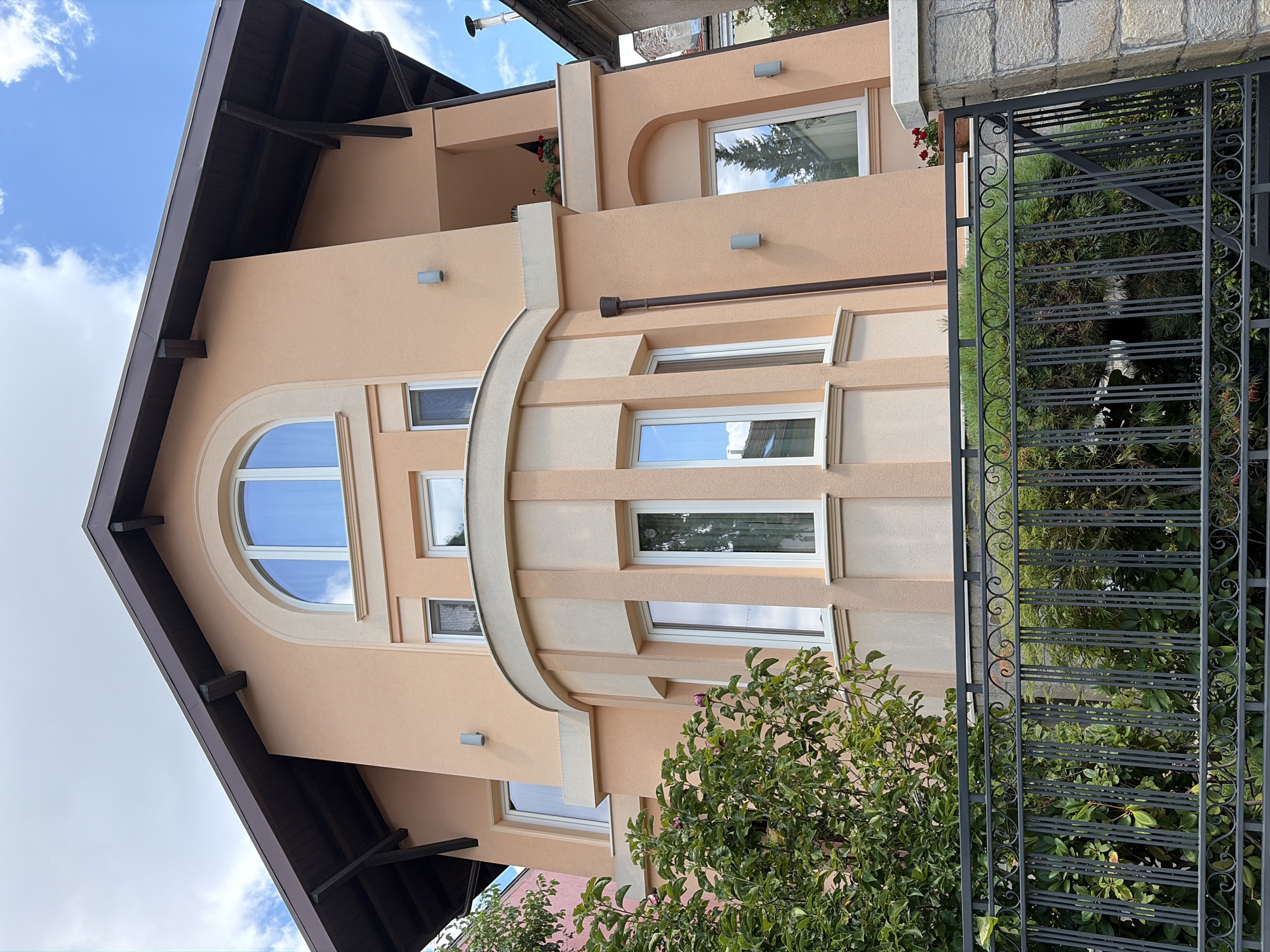 The image shows a two-story residential building with a peach-colored stucco exterior and a dark brown pitched roof with exposed wooden beams. The building features a curved bay window on the first floor with vertical reeded accents and arched windows on both floors. The bay window has multiple panes, and there are several other windows visible on the facade, most of which are rectangular with white frames. Two wall-mounted light fixtures are visible on the peach-colored walls. A small balcony with a railing is located on the second floor, with potted plants and flowers. In the foreground, a black wrought iron fence with decorative scrollwork separates the property from the street. Behind the fence, lush green foliage and trees are visible. The sky above is blue with scattered white clouds. The scene appears to be in a residential neighborhood in Sofia, Bulgaria, based on contextual information. There are no people visible in the image.