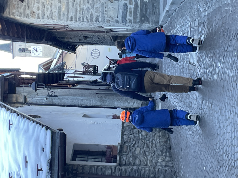 A group of people dressed in winter ski attire walks along a narrow, cobbled street in the mountain town of Courmayeur, Italy. The scene depicts a typical European alpine village, with buildings constructed from stone and plaster, some featuring traditional wooden balconies. Snow-covered rooftops are visible, confirming a cold, wintery environment.
In the foreground, a child wearing a bright blue ski suit and an orange helmet holds a ski pole. Behind them, an adult, dressed in a dark jacket and brown pants, carries a black backpack and also holds a ski pole. Further back, another child in a blue ski suit and ski boots, and a person in a red jacket, are part of the group. They appear to be either en route to or returning from skiing or snowboarding activities.
The street itself is paved with cobblestones, which appear slightly wet, reflecting the light. The time of day is bright, suggesting late morning or early afternoon. In the background, a clock tower with Roman numerals can be seen, with the clock face indicating approximately 10:50 or 10:55. The architectural style, winter conditions, and the subjects' clothing all reinforce the setting of a ski resort town.