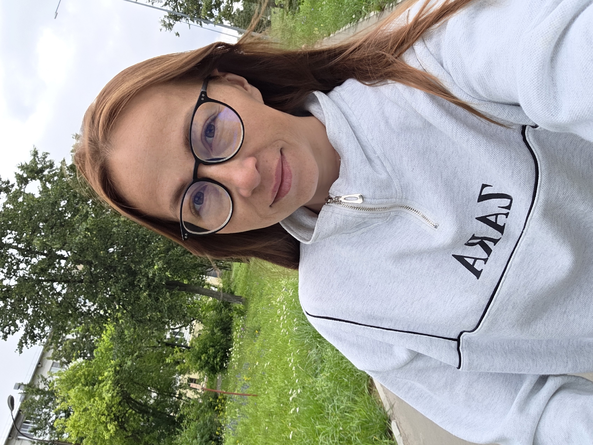 A woman with long auburn hair and glasses is taking a selfie outdoors. She is wearing a light gray sweatshirt with black trim and a zipper at the neck. Black text, possibly a logo, is visible on the front of the sweatshirt. She is standing on a paved pathway, with a grassy area dotted with wildflowers and trees behind her. In the background, a building and a street lamp are visible, consistent with an urban setting. The location is identified as Chaikovsky, Russia. The lighting suggests it is daytime, with overcast skies.