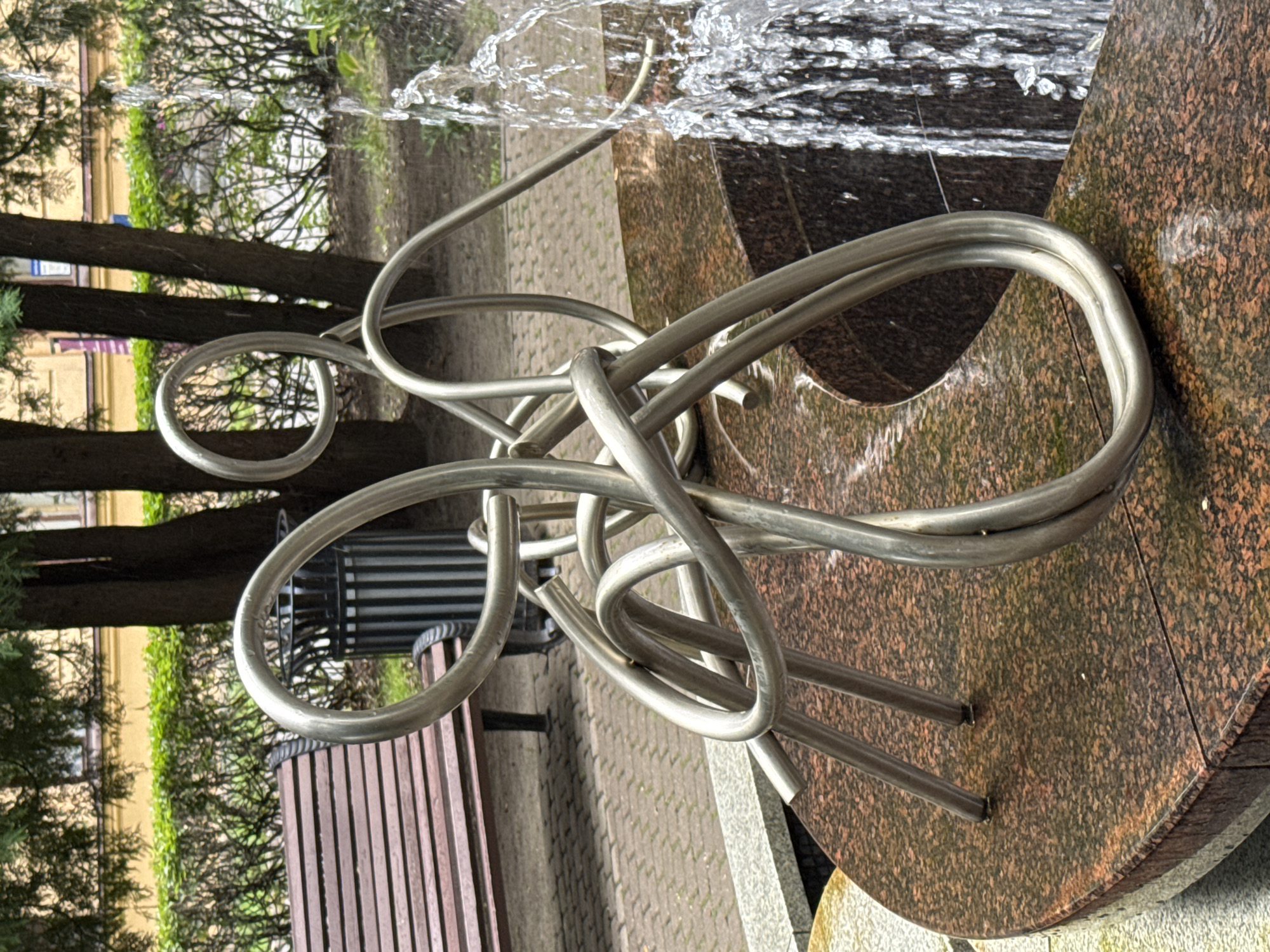 The image shows a metallic sculpture resembling a tangle of wires, positioned on the edge of a granite fountain in what appears to be a park or public square. The sculpture is made of bent and twisted silver-colored metal tubes, forming abstract shapes. To the left of the sculpture, a wooden park bench with metal supports is visible, and behind that, a trash can with vertical slats. The background features trees with green foliage and a building with a yellow facade. A fountain is active on the right side of the image, with water spraying upwards and cascading down. The ground in front of the bench is paved with interlocking bricks. The overall scene suggests an outdoor urban setting, possibly a pedestrian area or a small garden within a city. Given the architecture visible and the style of the urban elements, this scene could be located in Saint Petersburg, Russia.