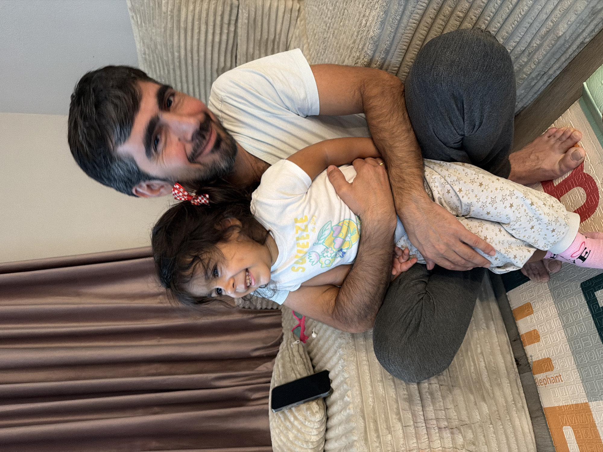An adult male and a young child are on a sofa. The male is wearing a white t-shirt and gray sweatpants. The child is wearing a white t-shirt with a yellow graphic and patterned pants. Both are smiling and looking in the same direction. The male is holding the child. A phone, a pillow, and a curtain are visible in the background. This image was taken indoors, likely in a residential setting. No specific location within Phoenix, United States of America can be determined from the provided image.