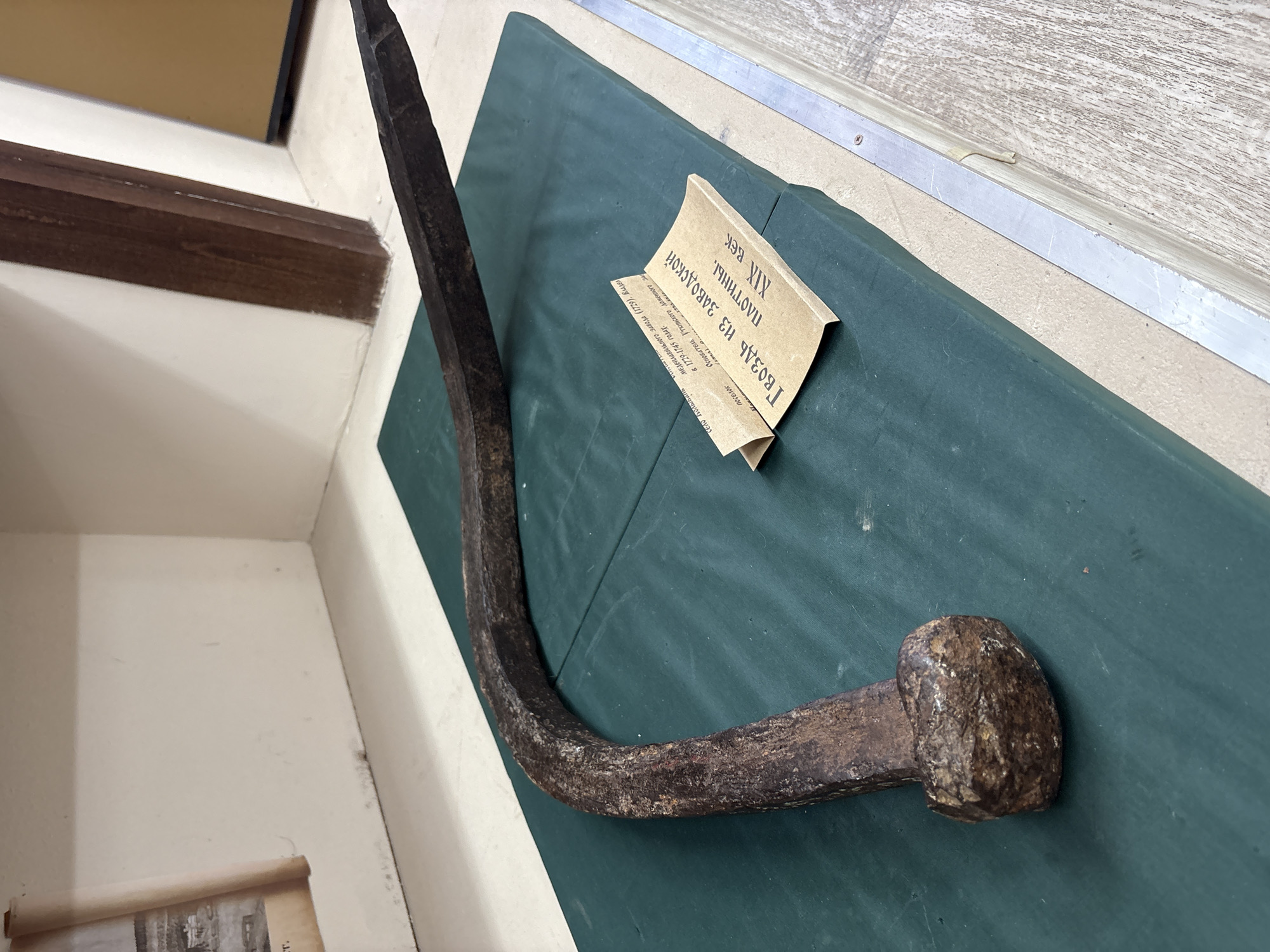 The image shows a large, curved, rusty nail exhibited on a dark green surface, possibly a display mat. To the right of the nail, a folded piece of light brown paper is placed, serving as a label. The label contains Cyrillic text and appears to be an informational card about the artifact. The background suggests an indoor setting, possibly a museum or historical exhibition, with a light-colored wall and wooden elements visible. The nail is the central subject, its substantial size and aged appearance suggesting historical significance.
