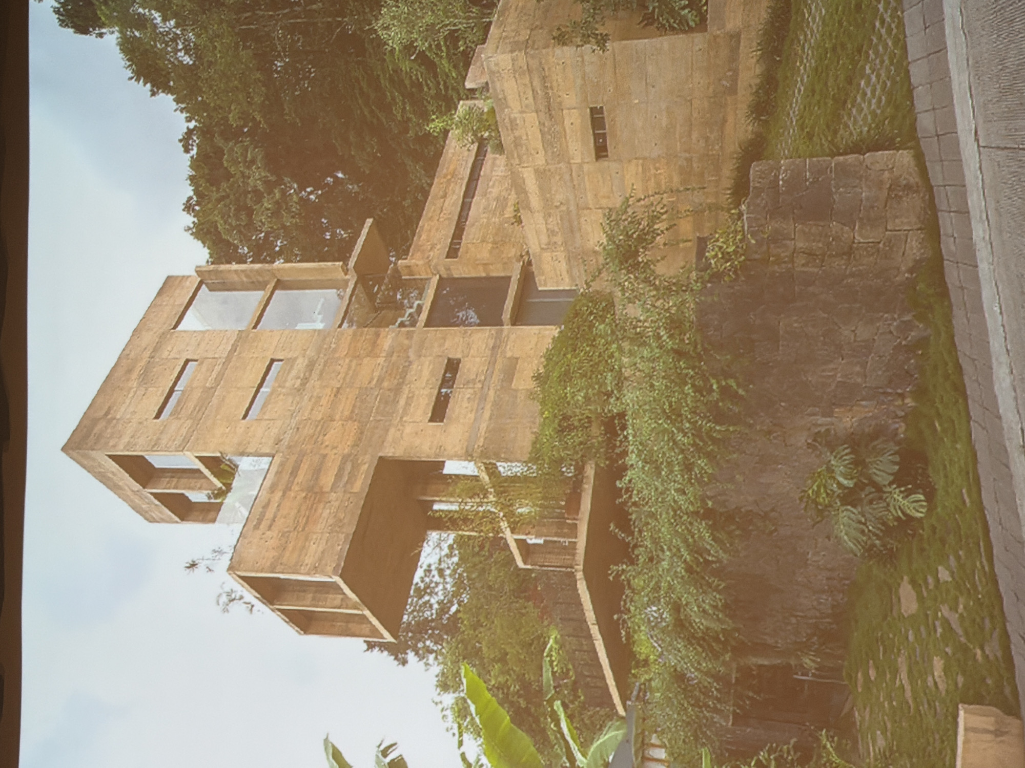 The image shows a concrete building set in a lush, green environment. The building has a geometric, modular design with open windows and vertical design of concrete slabs. The surrounding vegetation is dense with various plants and trees, creating a natural backdrop. The building appears to be well-integrated into the landscape.