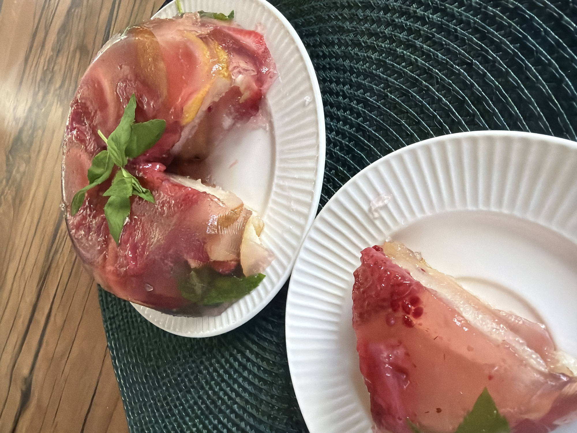 A close-up, eye-level shot captures a partially eaten fruit gelatin cake on a white plate, which is placed on a dark green woven placemat. The cake, which appears to be a dome shape, is layered with what look like strawberries, lemons, and possibly other fruits, suspended in a clear gelatin. A few fresh green leaves, possibly mint or basil, are placed on top of the cake as garnish. To the left of the cake, a slice has been removed and is presented on a separate, smaller white plate. The slice reveals the layered interior of the gelatin and the embedded fruits. The background consists of a wooden table with a prominent grain pattern. The lighting is natural, casting soft shadows and highlighting the translucency of the gelatin. The scene is static and domestic, with no individuals present. The location in the city of Steklyannyi, Russia, is an unconfirmed detail and not visually supported by the image content.