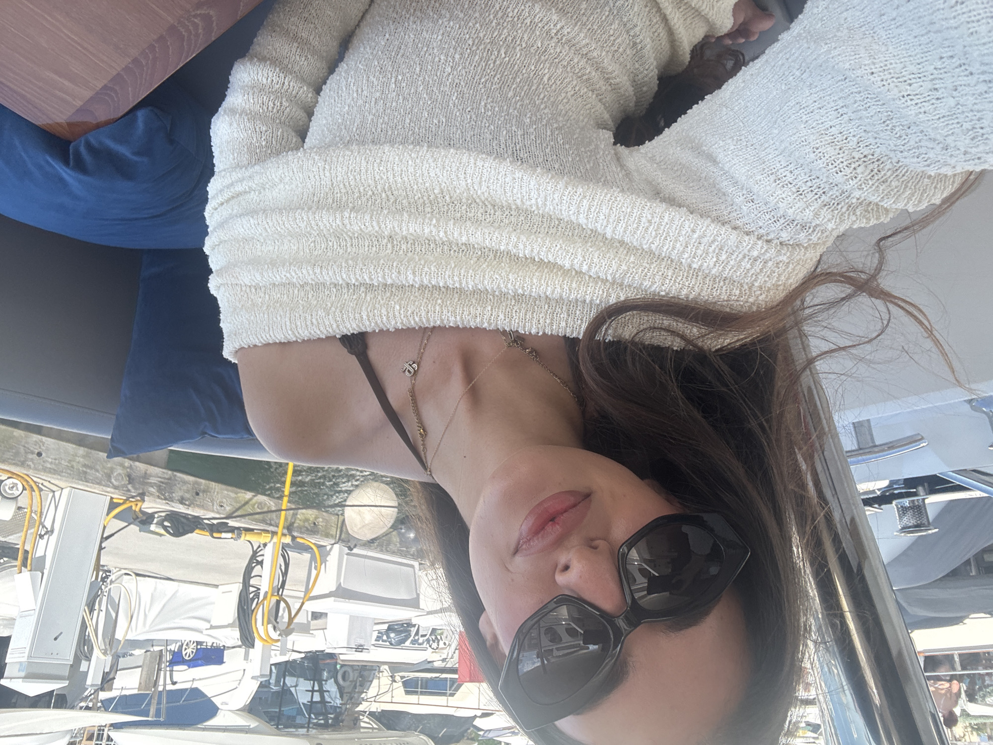 A woman wearing a cream-colored, textured sweater and dark sunglasses is the central subject of the image. She is also wearing gold necklaces. The background shows a marina setting, with numerous boats docked. Specific details visible include the boats' equipment, wiring, and the pier. While the image provides a glimpse of boats and a dock, the precise location within Miami Beach cannot be determined from the provided visual data.
