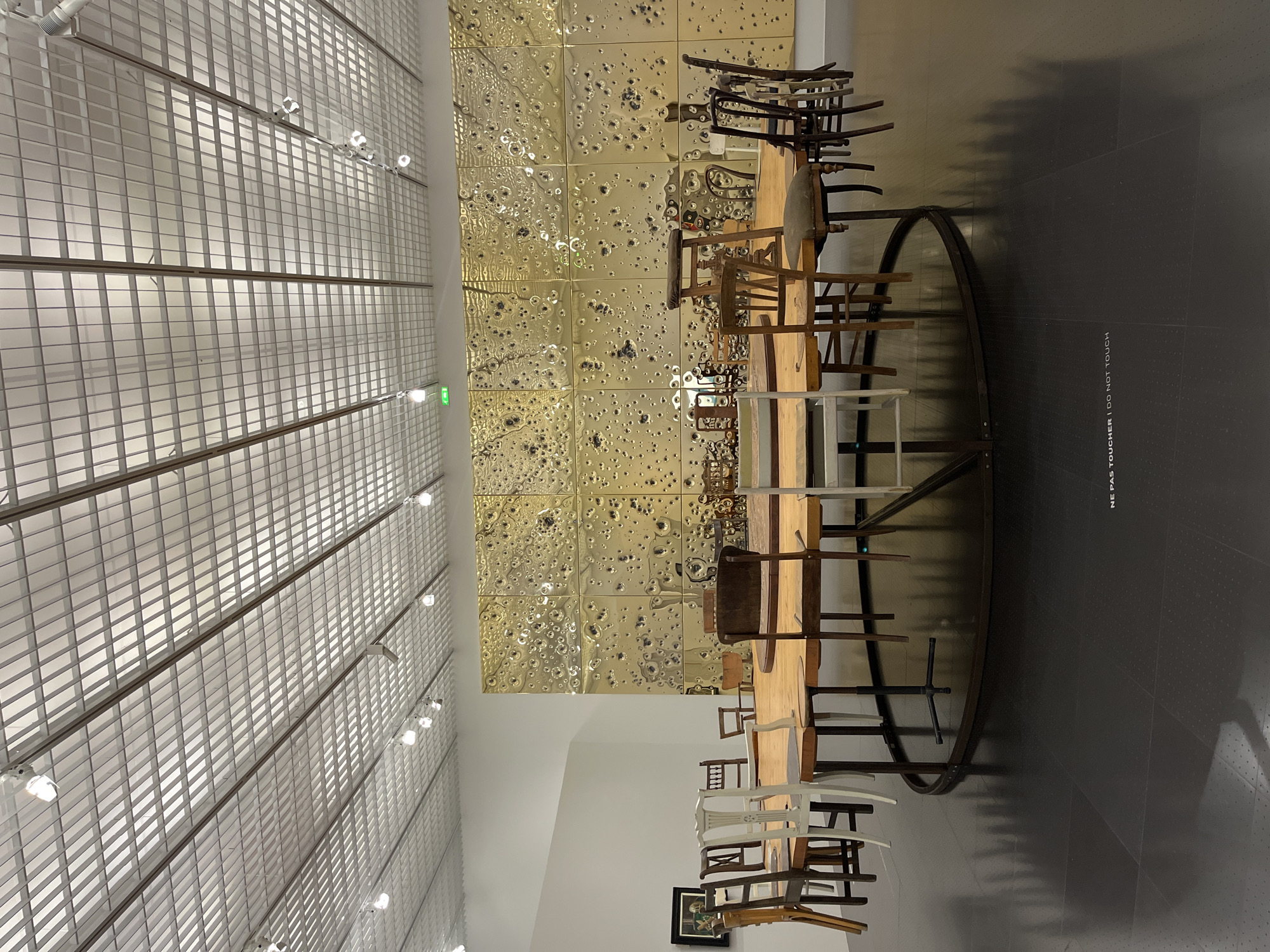 The photograph depicts an art installation at the Centre Pompidou-Metz in Metz, France. The installation features a circular table surrounded by a variety of chairs, arranged in a ring. The backdrop is a gold-colored wall with a pattern of round indentations or bumps. Overhead, there is a grid-like ceiling structure with integrated lighting. On the right side of the image, the floor is dark gray. A sign with text that says “NE PAS TOUCHER / DO NOT TOUCH” is visible on the floor.