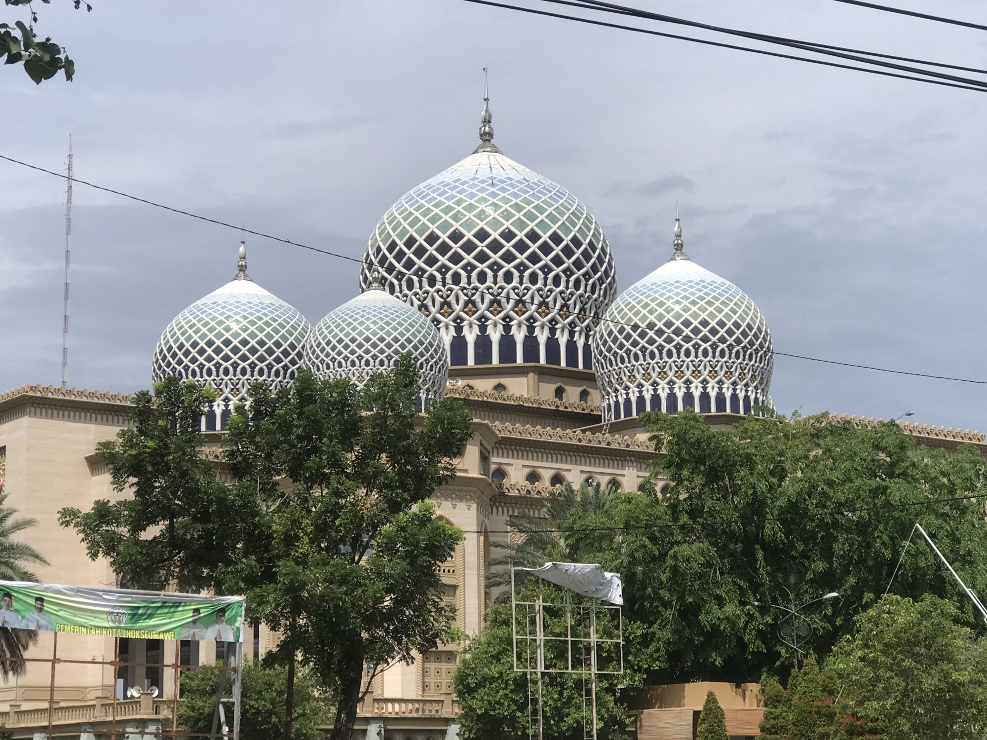 The image depicts a building with multiple domes, partially obscured by trees. The largest dome is in the center, with three smaller domes flanking it. The domes are decorated with a geometric pattern, and the building's facade is ornate. A banner with Indonesian text and images of people is visible in the foreground, identified as "PEMERINTAH KOTA LHOKSEUMAWE," indicating the location is Lhokseumawe, Indonesia. A tall antenna is visible in the background, on the left side of the image. Power lines stretch across the top of the frame. The sky is overcast, suggesting a cloudy day.