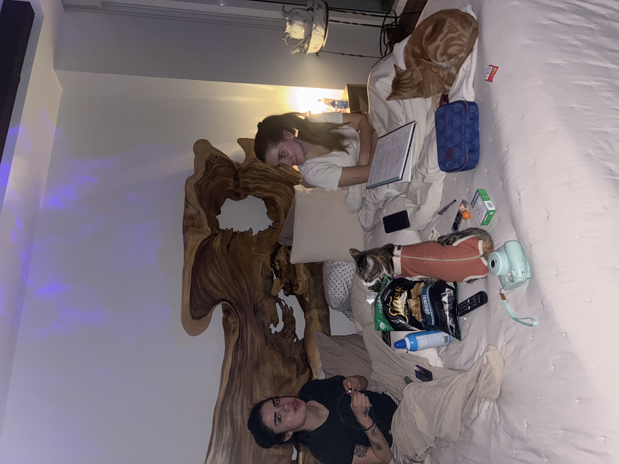 A bedroom scene in Huixquilucan de Degollado, Mexico shows two women and two cats on a large bed with a prominent wooden headboard. One woman with dark hair is lying on her back at the bottom of the bed, wearing a black shirt, with a blanket partially covering her. The other woman, with lighter hair pulled back in a ponytail, is propped up in the middle of the bed, holding a notebook. An orange cat is curled up on the top right corner of the bed. A smaller cat wearing an orange sweater is lying near the middle of the bed. Various objects are scattered on the bed, including a blue makeup bag, a camera, a bag of chips, a TV remote, and a bottle of lotion. The room is lit by a bedside lamp.