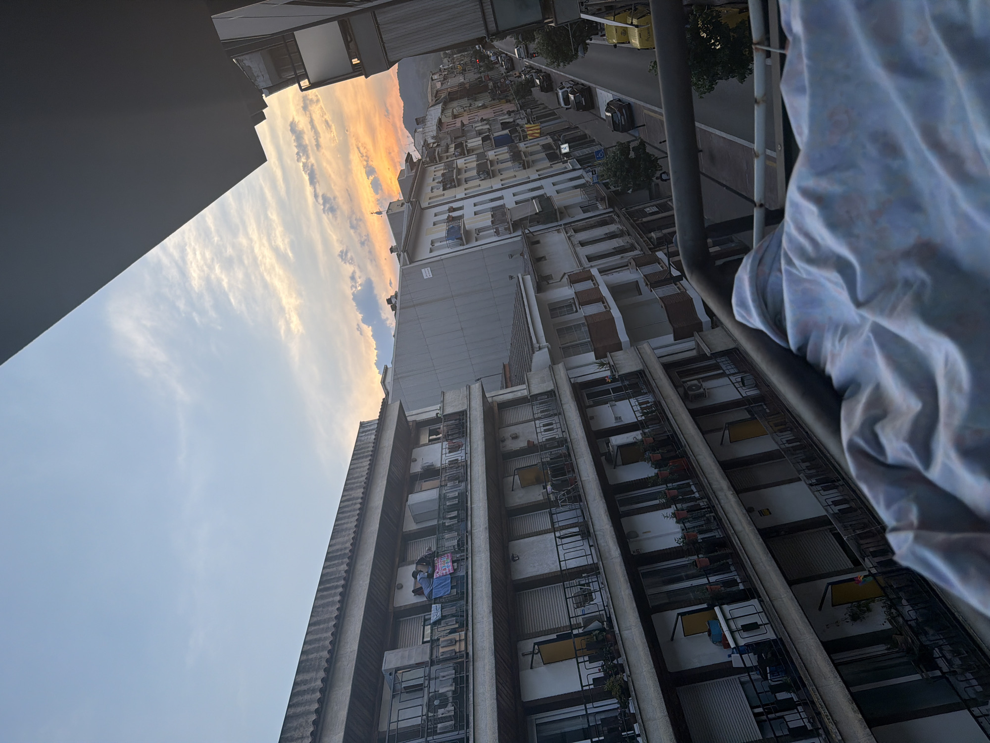 The image captures a view from a balcony overlooking a street in Arbúcies, Spain, during sunset. The sky displays hues of orange and yellow due to the setting sun, with scattered clouds. In the foreground, a crumpled blanket is visible, covering a clothes drying rack.
The main subject of the image is a multi-story building with balconies, common in urban European settings. Several balconies are occupied by various items. On one of the upper balconies, two individuals are standing, and laundry is hanging. Other balconies have plants and shutters.
Below the buildings, a street runs through the town, with several cars parked or driving on it. The architecture of the surrounding buildings is typical of a Spanish town, with balconies and multiple floors. Trees line the street, adding greenery to the urban landscape. In the distance, hills or mountains are visible, silhouetted against the colorful sky. The overall impression is a peaceful, everyday scene in a Spanish town at dusk.