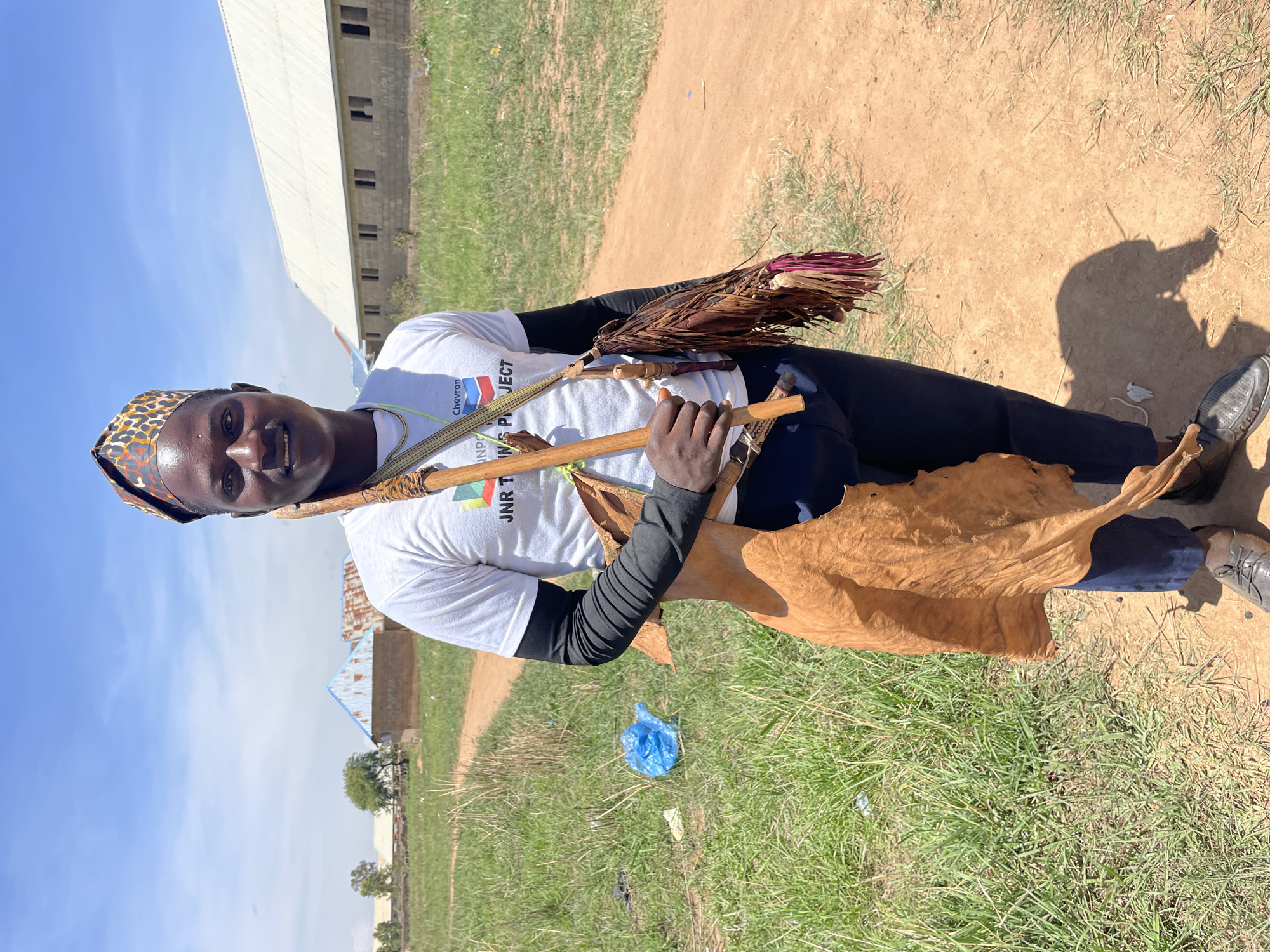 A person wearing a patterned head covering, a white t-shirt with visible logos, and black long-sleeved underclothes is standing in an outdoor area. They hold various objects: a bundle of dry leaves, wooden sticks, and a large piece of what appears to be animal hide. The ground is a mix of green grass and bare dirt. In the background, there is a building, possibly a warehouse or industrial structure, and a couple of smaller structures. A blue plastic bag lies discarded on the grass. The location is identified as Durumi, Nigeria, although specific details that might pinpoint the exact location within the city are not readily apparent from the image.