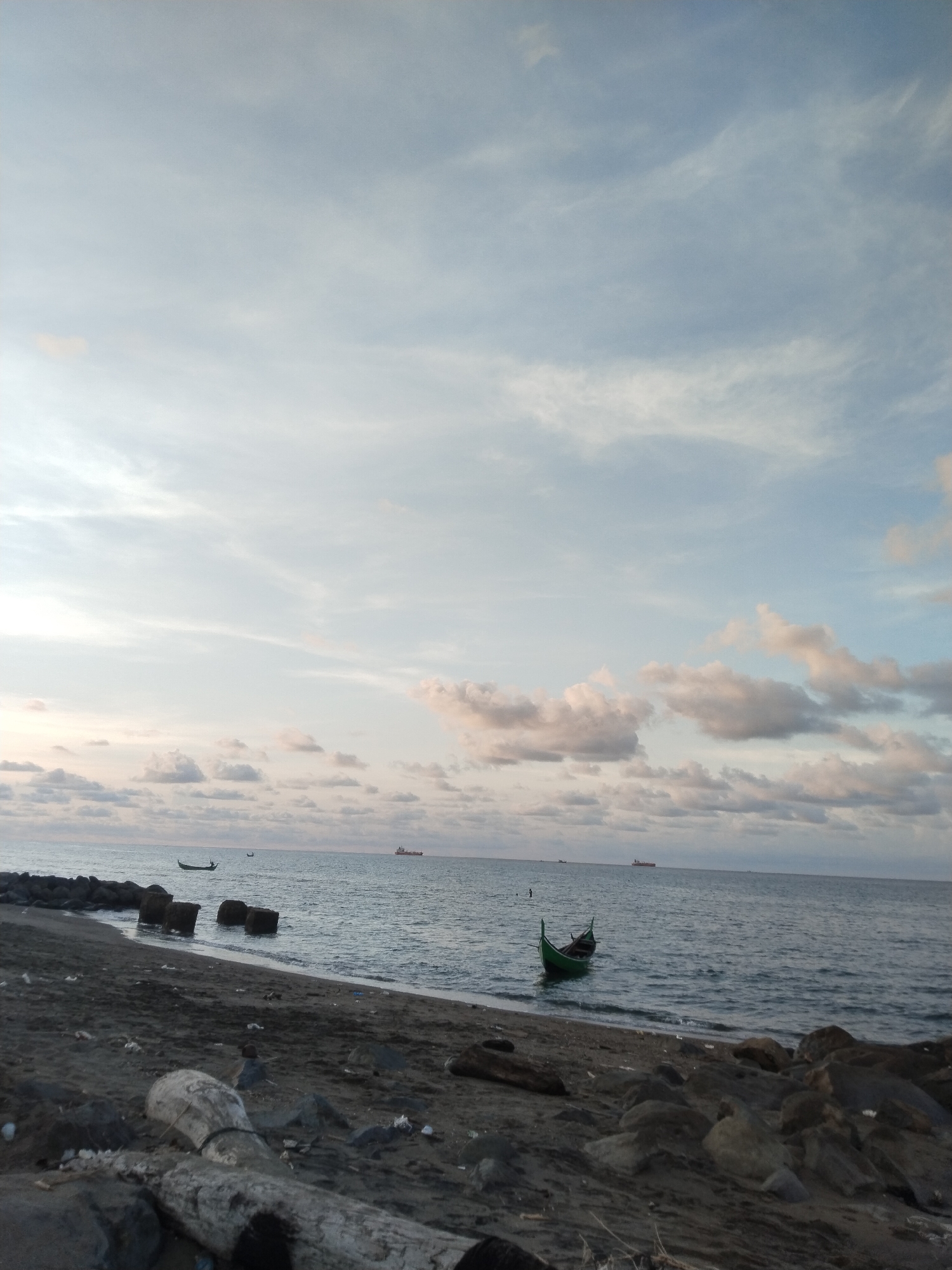 The image shows a beach in Lhokseumawe, Indonesia. The sky is overcast with light clouds. The sea is calm with several ships in the distance. A small, green boat is docked near the shore. The beach is rocky and littered with debris, including pieces of driftwood. No people are visible.